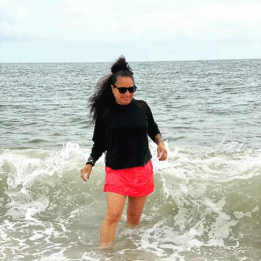 Lydia wearing a pink skirt and sunglasses standing in the ocean on a cloudy day