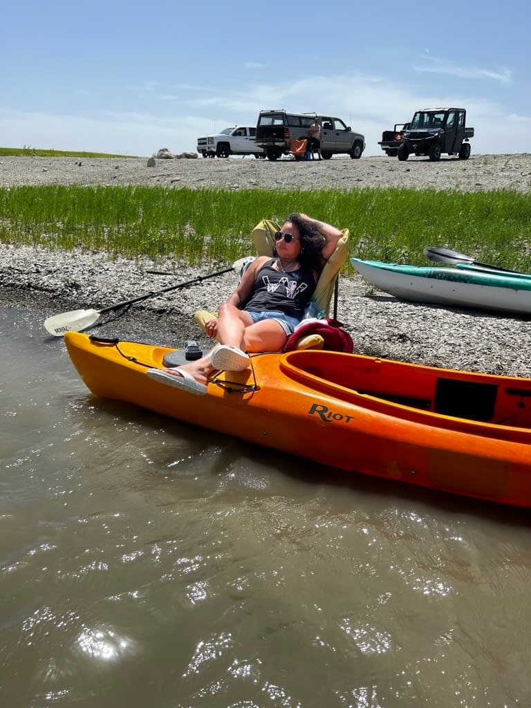 Lydia relaxing with her feet on a kayak on the bank of a river with cars in the background