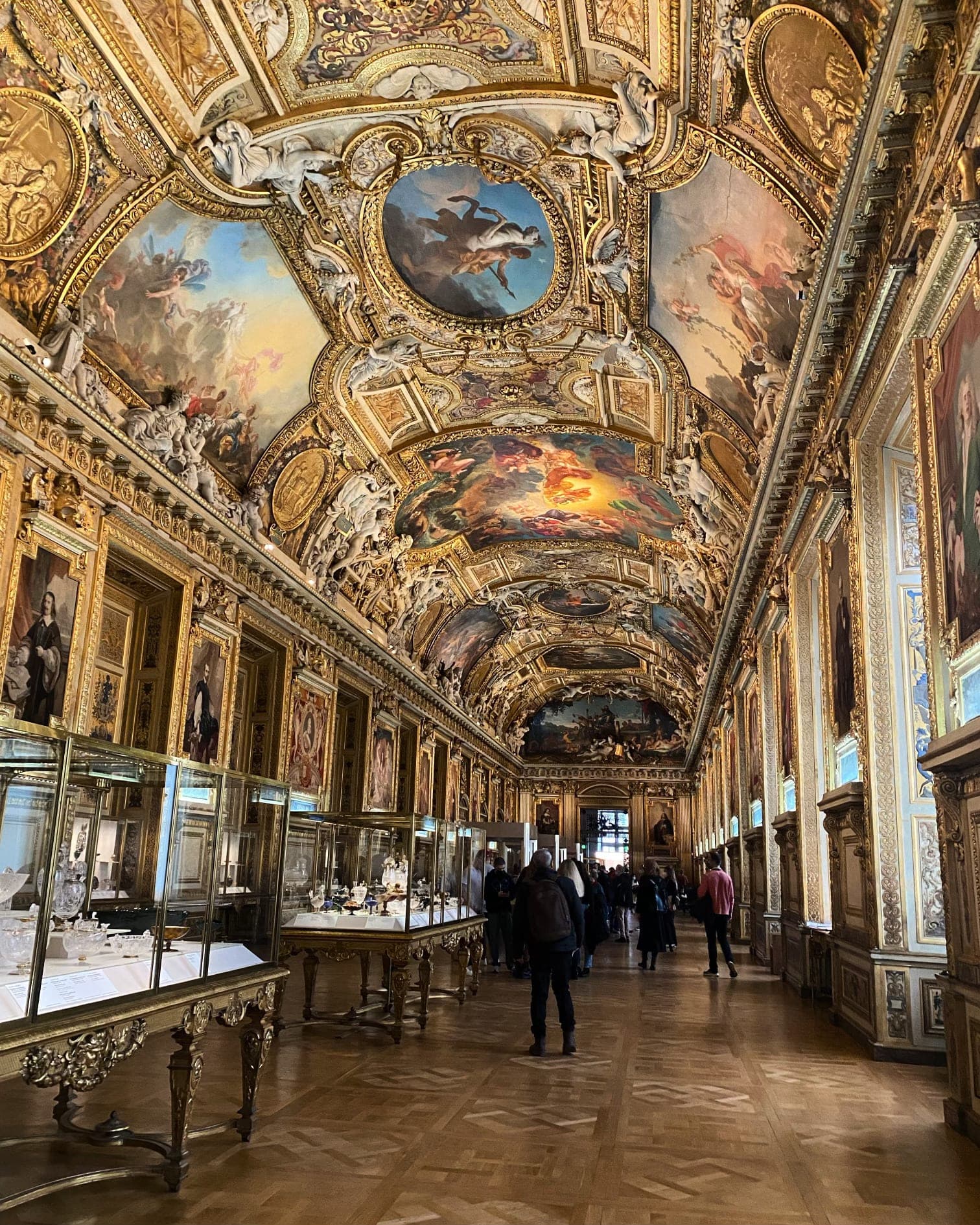 Mostly empty hall in the Louvre Museum featuring ornately painted ceilings and glass cases on display