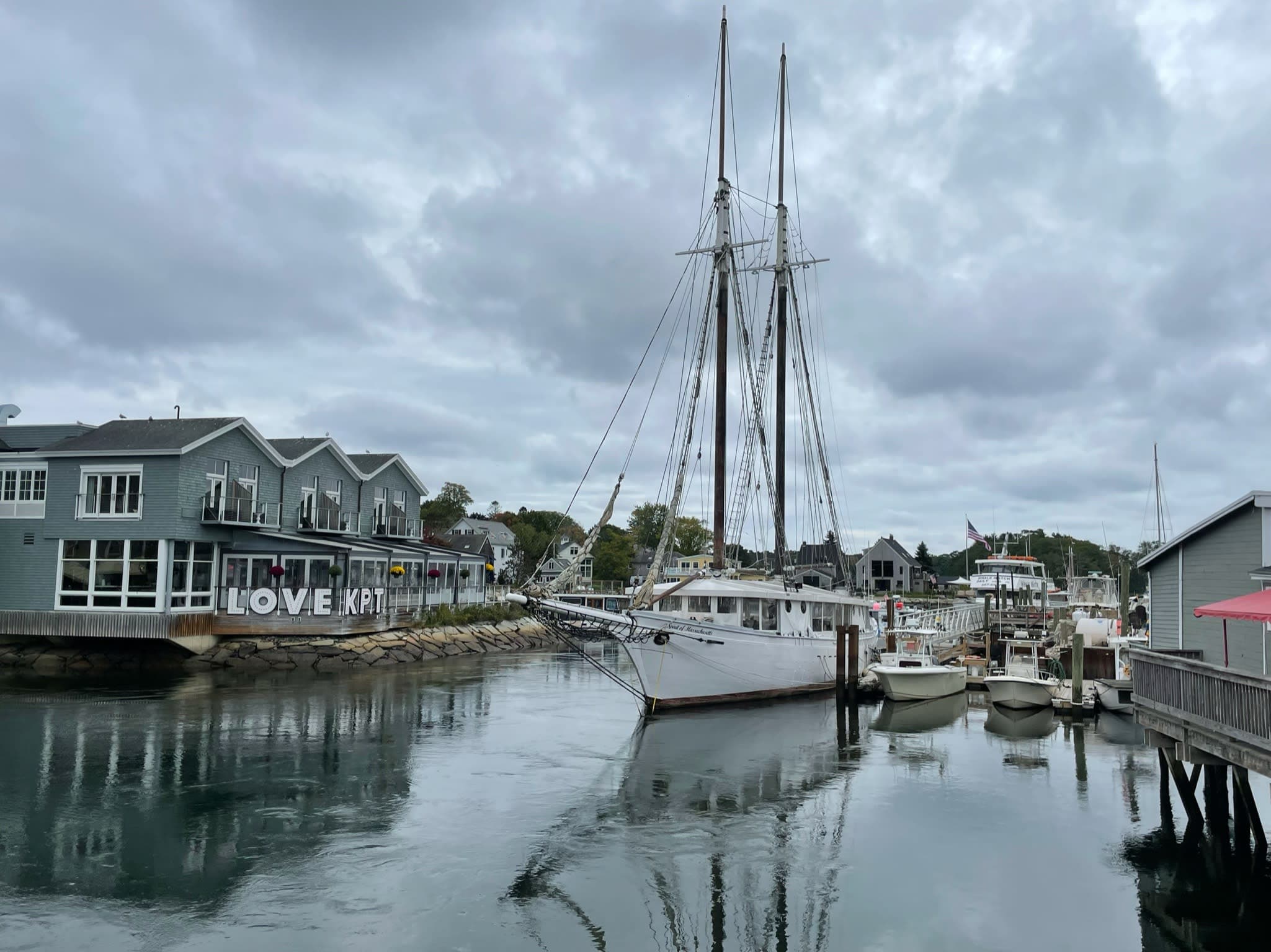 View of a large sailboat docked in a marina with small buildings on either side on a cloudy day