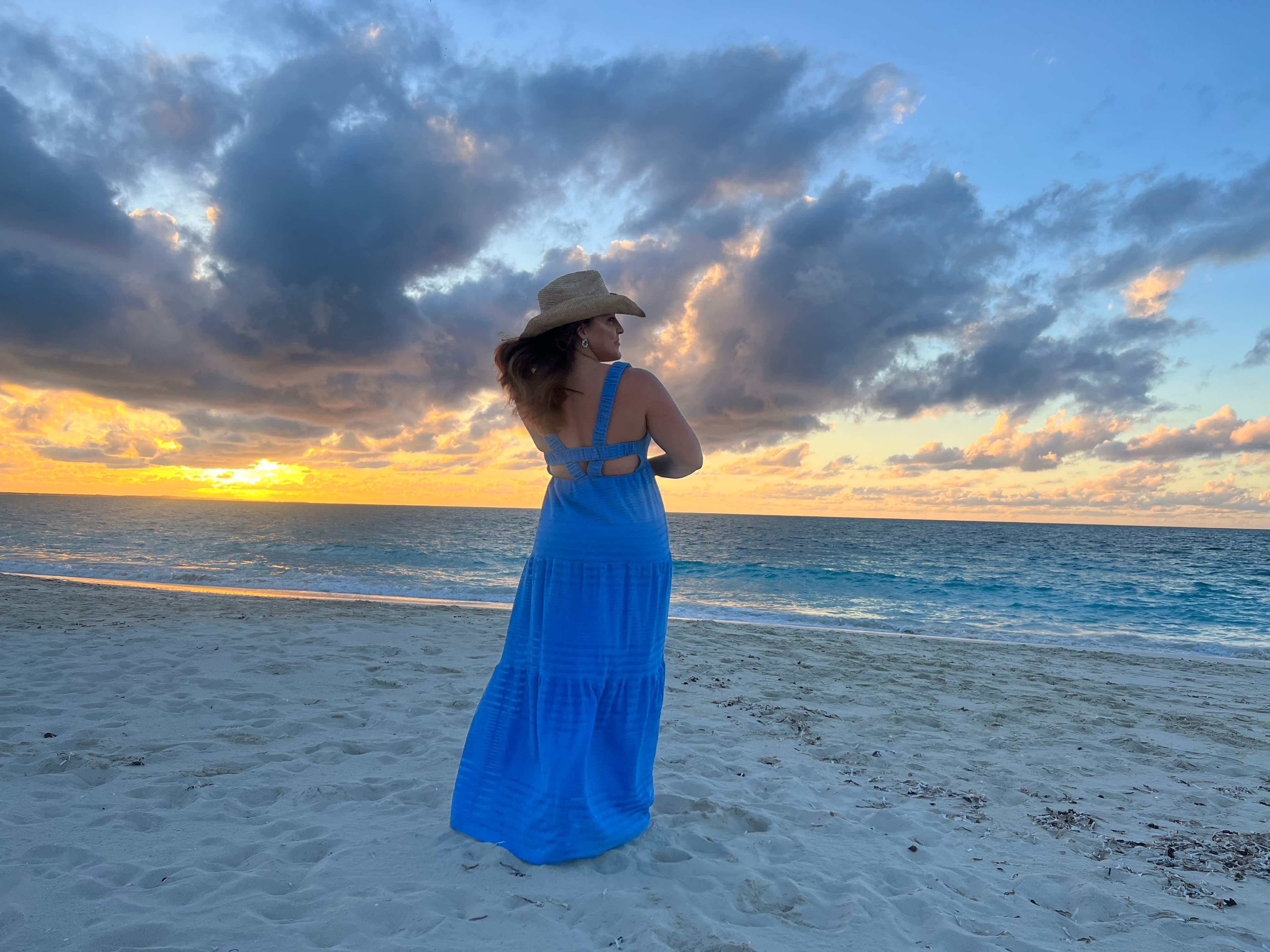 Leah, standing on a beach at sunset in a long blue dress and cowboy hat. 