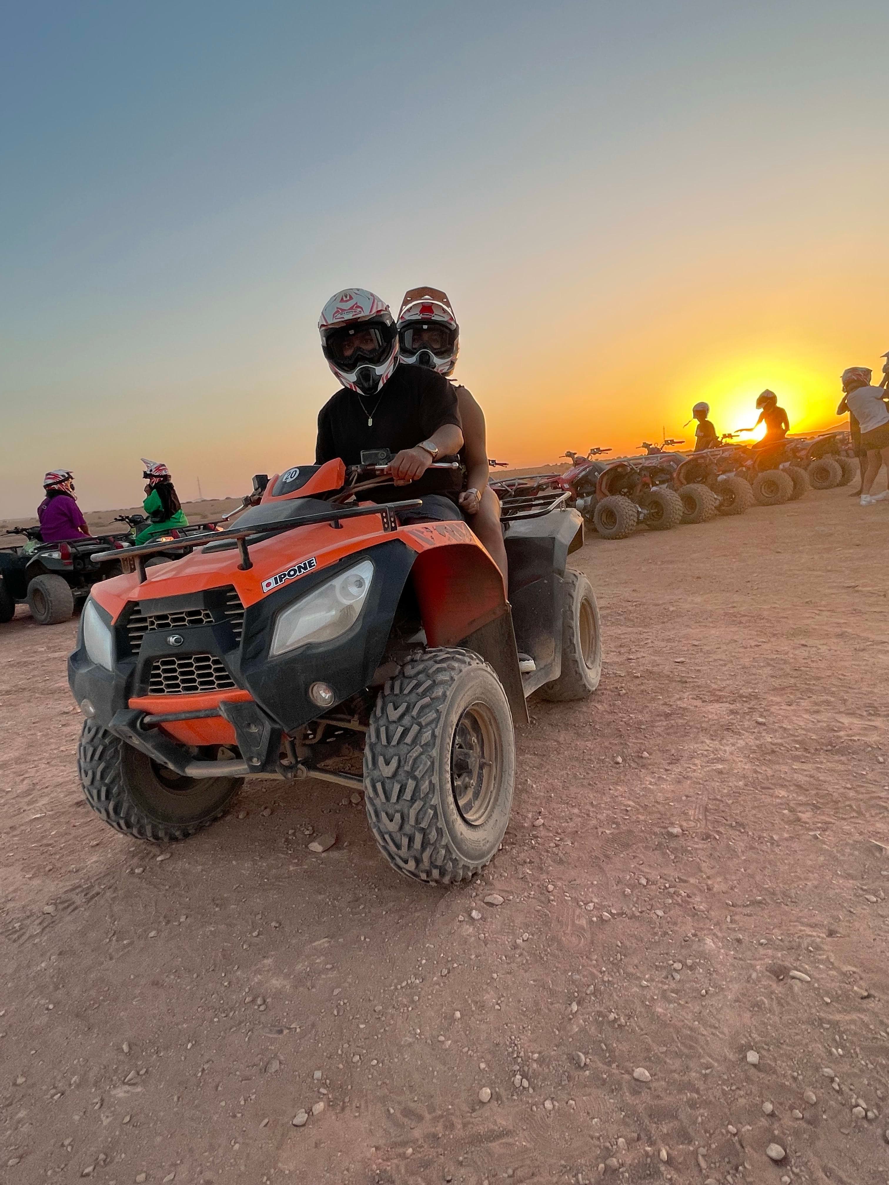 View of two people wearing helmets and riding a red ATV at sunset in the desert