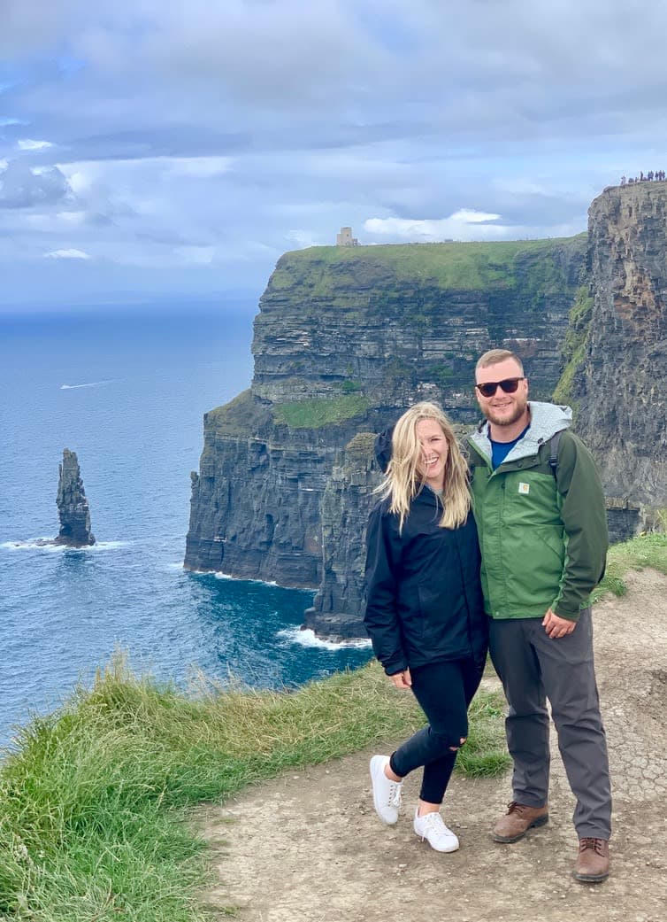 Olivia and her partner smiling in jackets along beautiful oceanside cliffs on a cloudy day