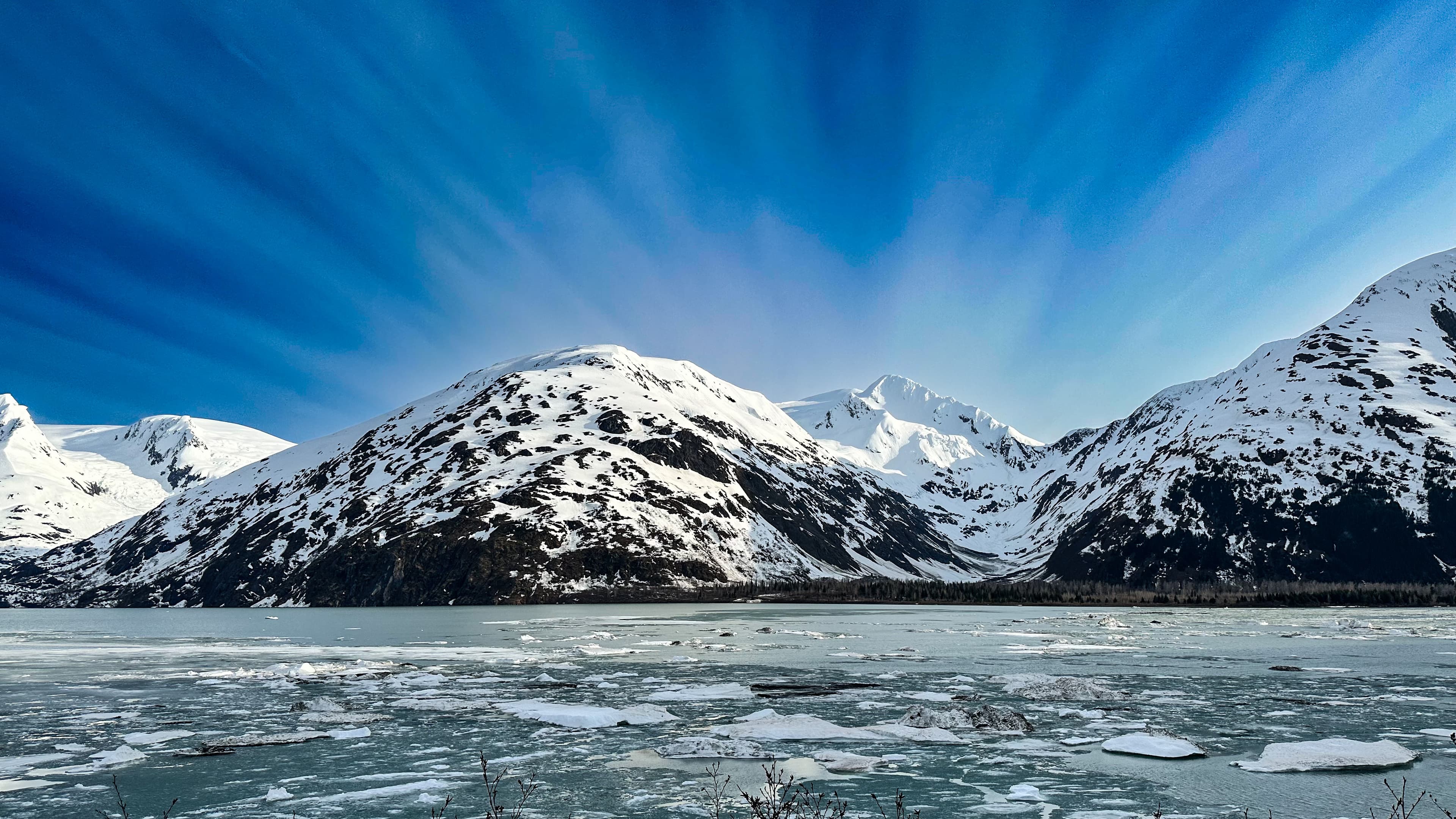 An icy water body with snow-capped mountains behind on a sunny day.