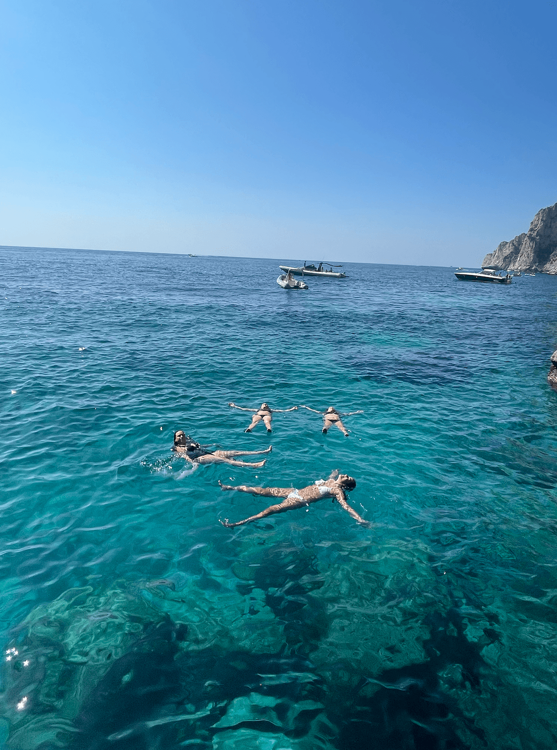 View of Sofia and friends floating on their backs in a crystal clear sea with small boats in the distance