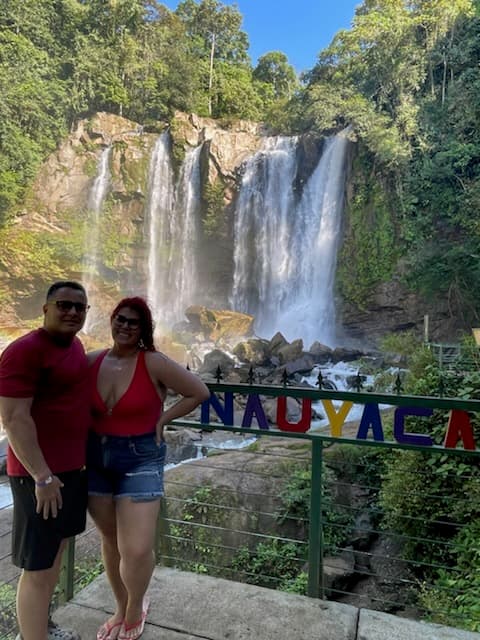 Advisor posing with her husband on a lookout point with a waterfall and foliage in the distance on a sunny day. 