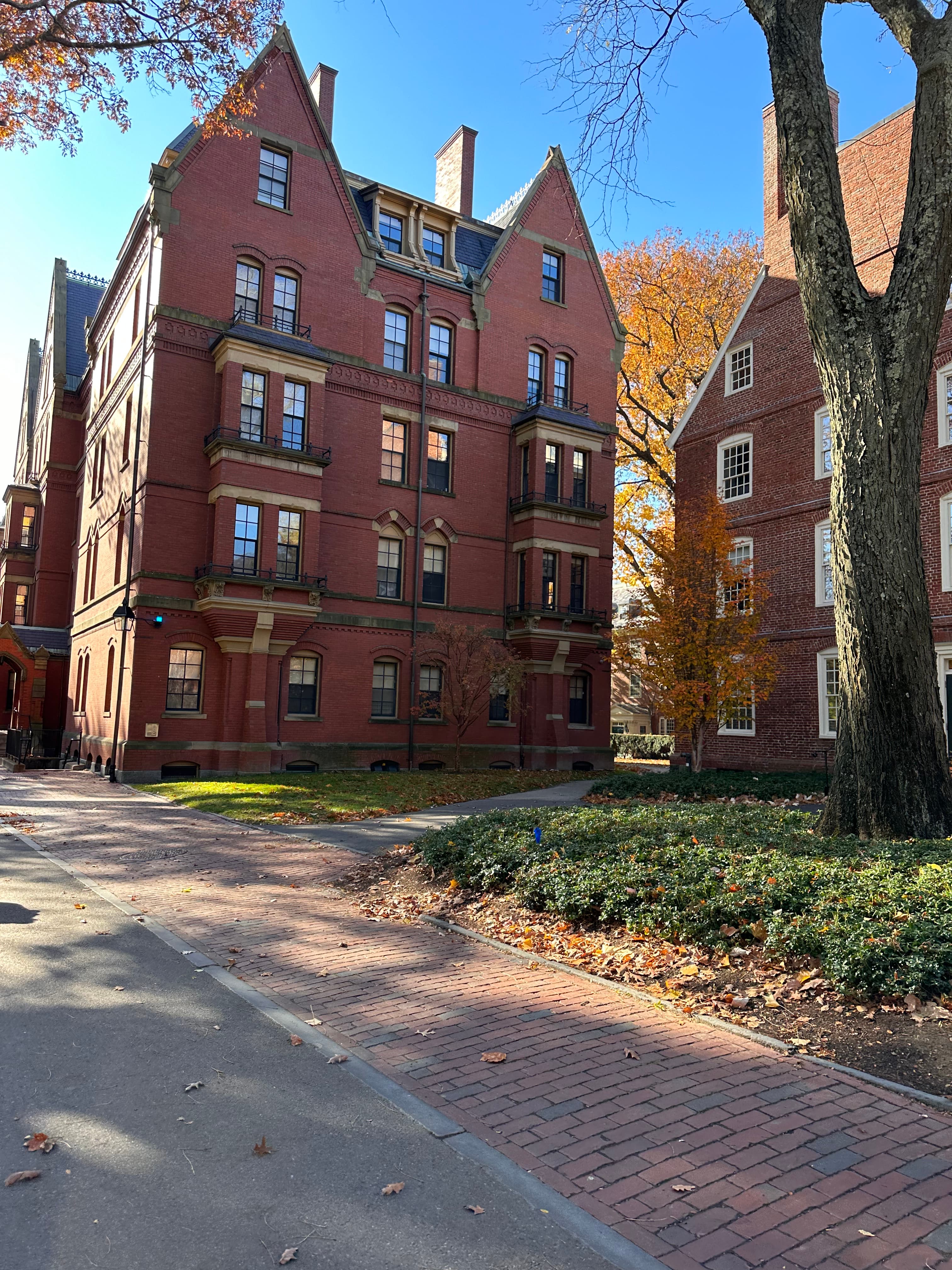 An old brick building with pointed roofs and a brick pathway leading up to it on a fall day. There are other buildings of a similar style behind it along with trees with orange leaves. 