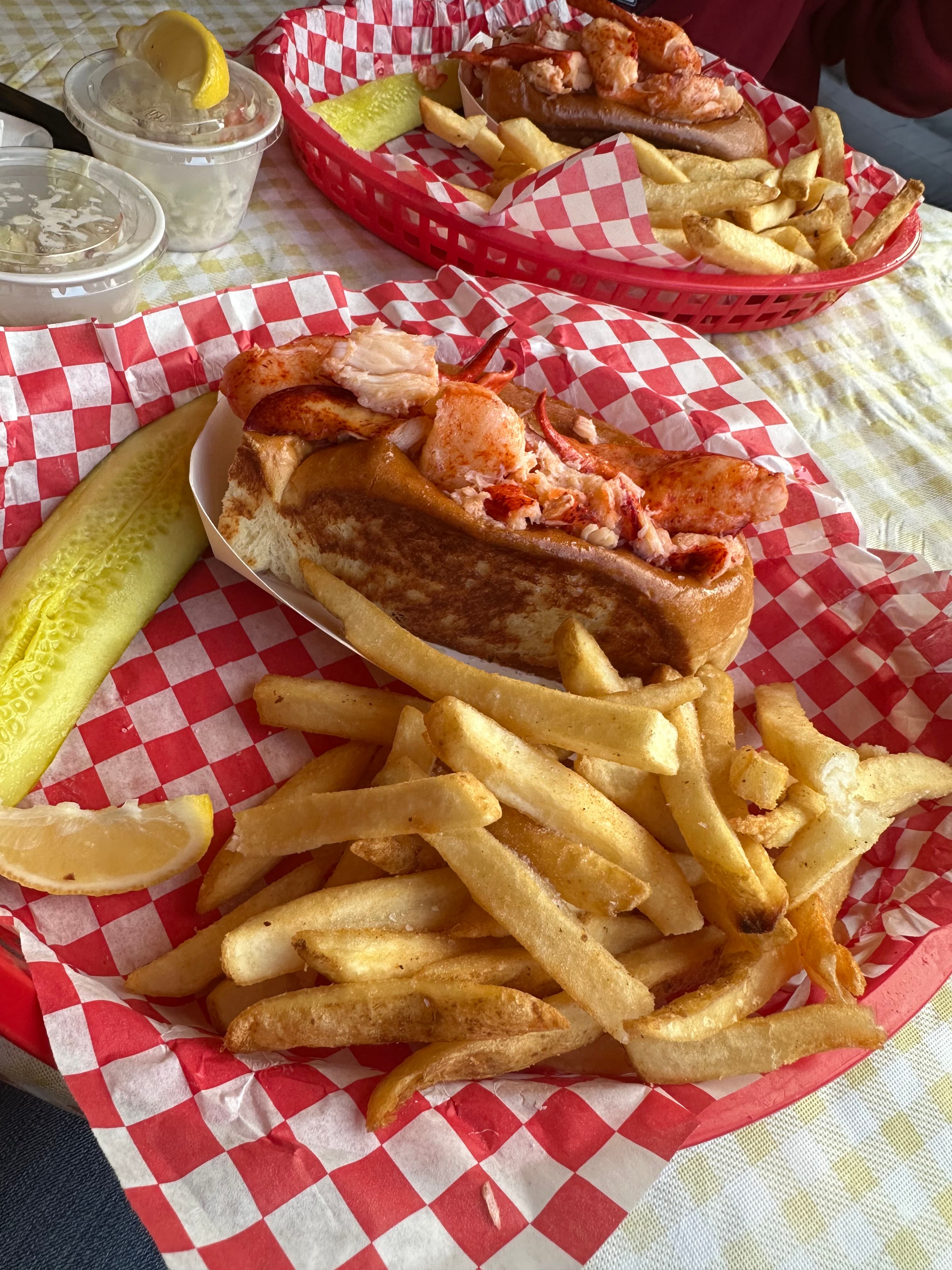 A diner-style plastic tray with red and white checkered liner holding a lobster roll, french fries, a pickle spear and a lemon wedge. 