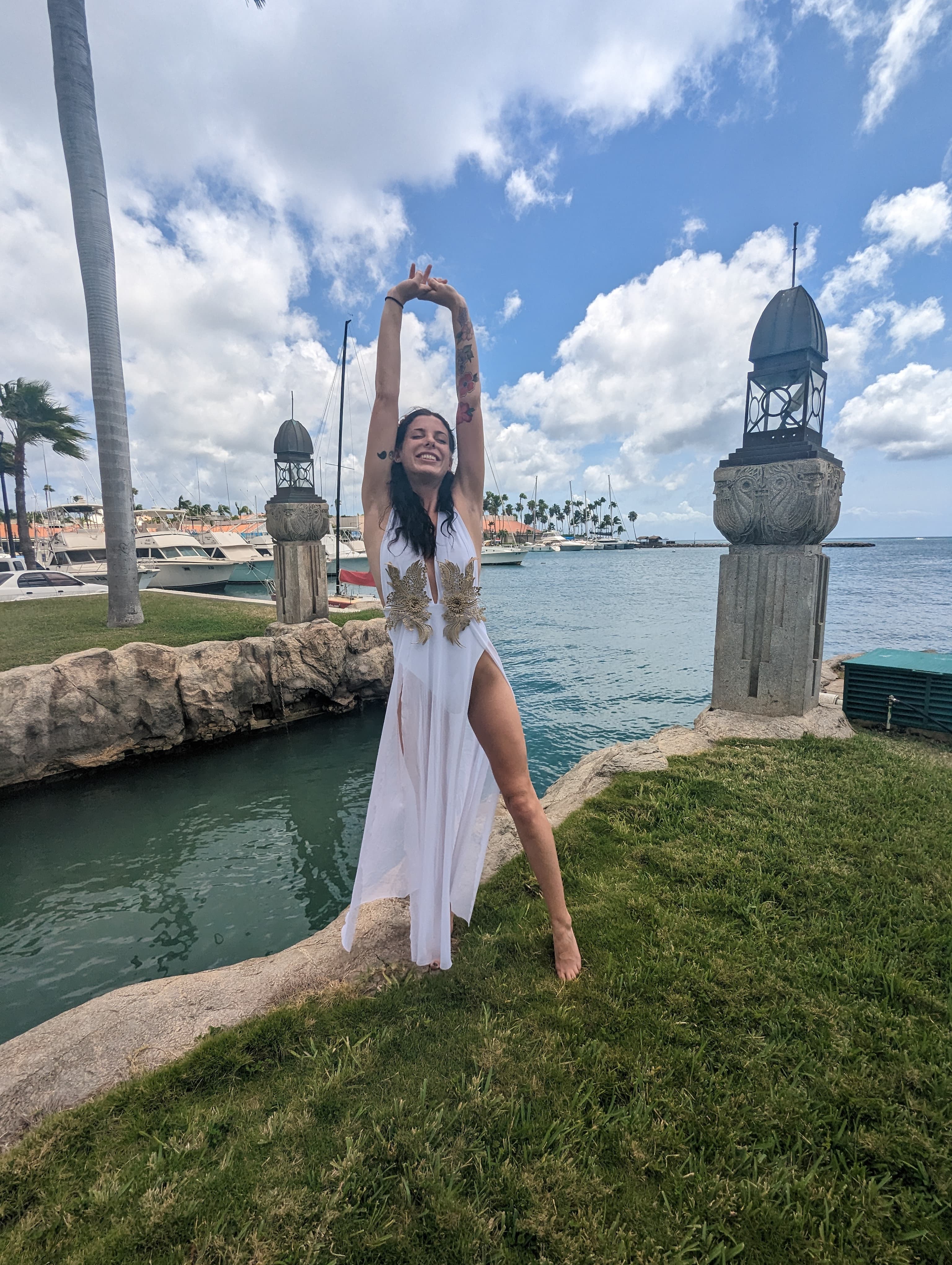 Image of Nicole smiling in a white dress with arms raised and bare feet standing in the grass along the sea