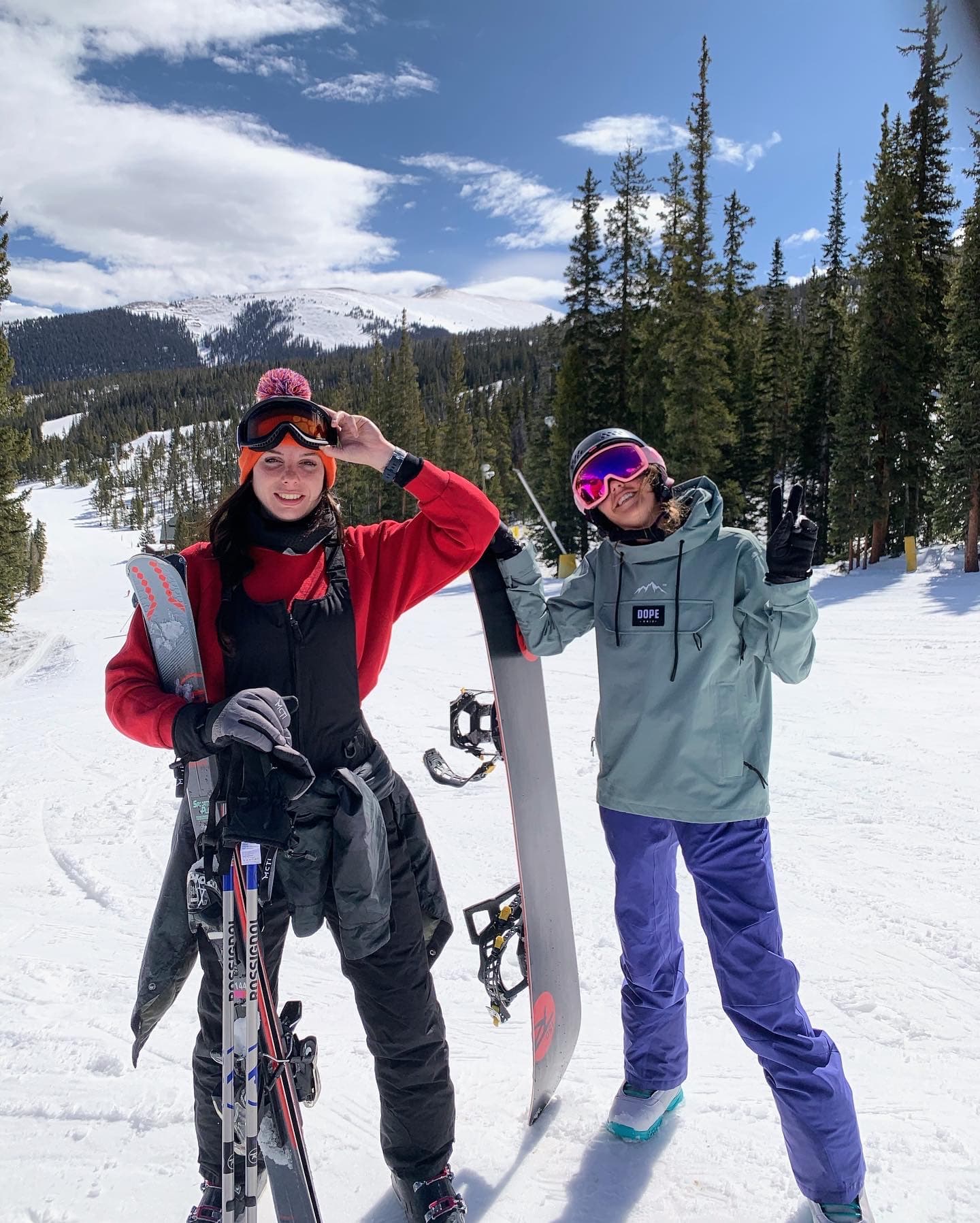 Nicole and a friend in winter jackets holding skis with a snowy landscape and tall pine trees in the background