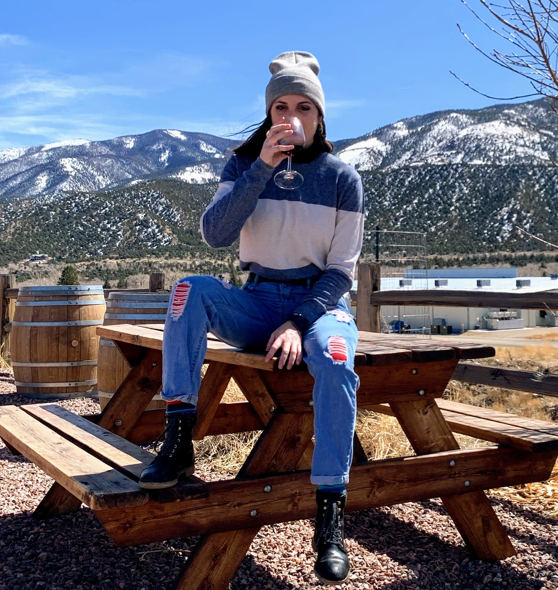 Nicole sitting on a picnic table wearing a beanie and holding a glass of wine with a stunning snowy mountain landscape behind her
