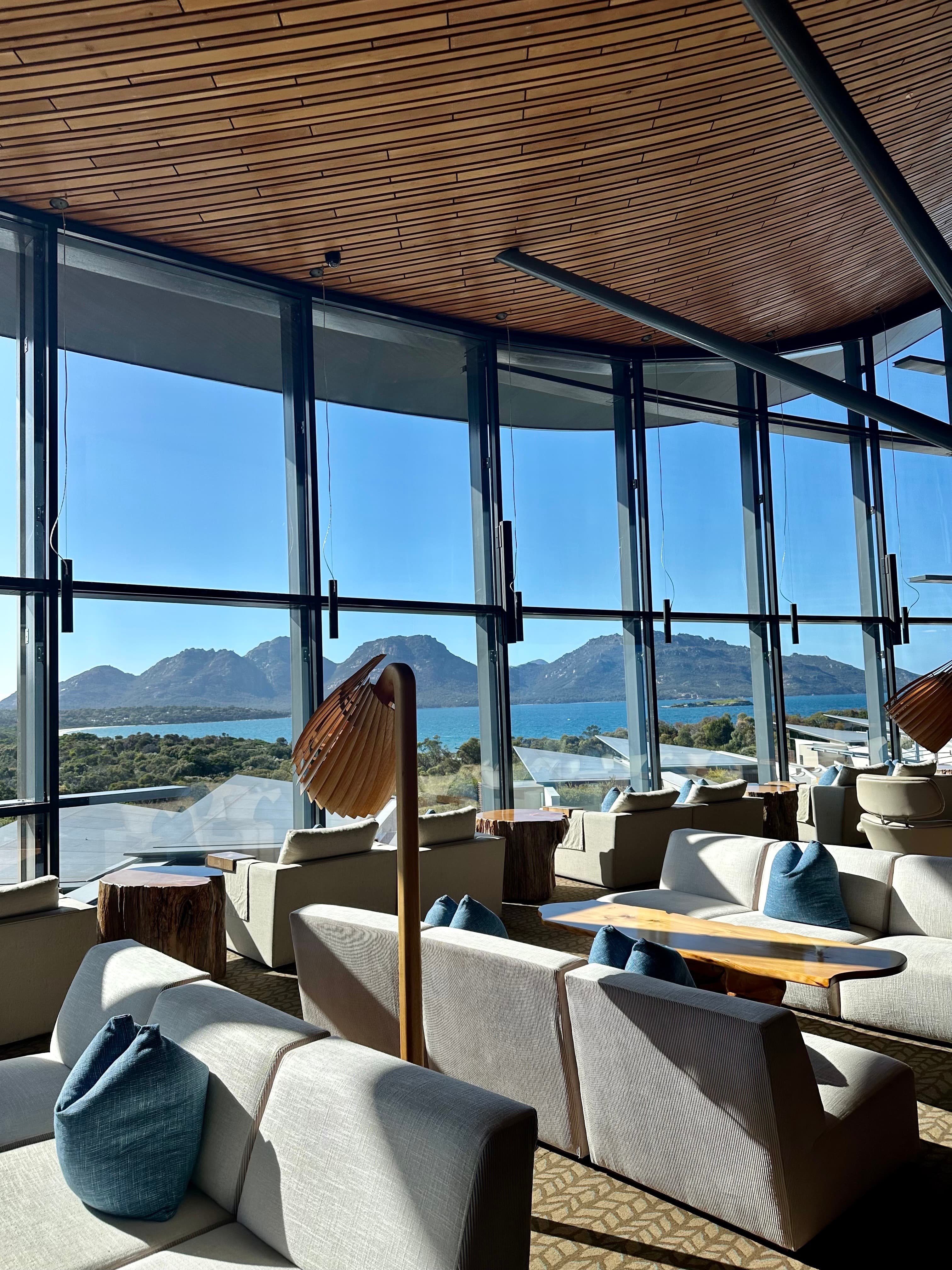 View of an indoor seating area with several couches and large glass windows looking towards beautiful water and mountain scenery