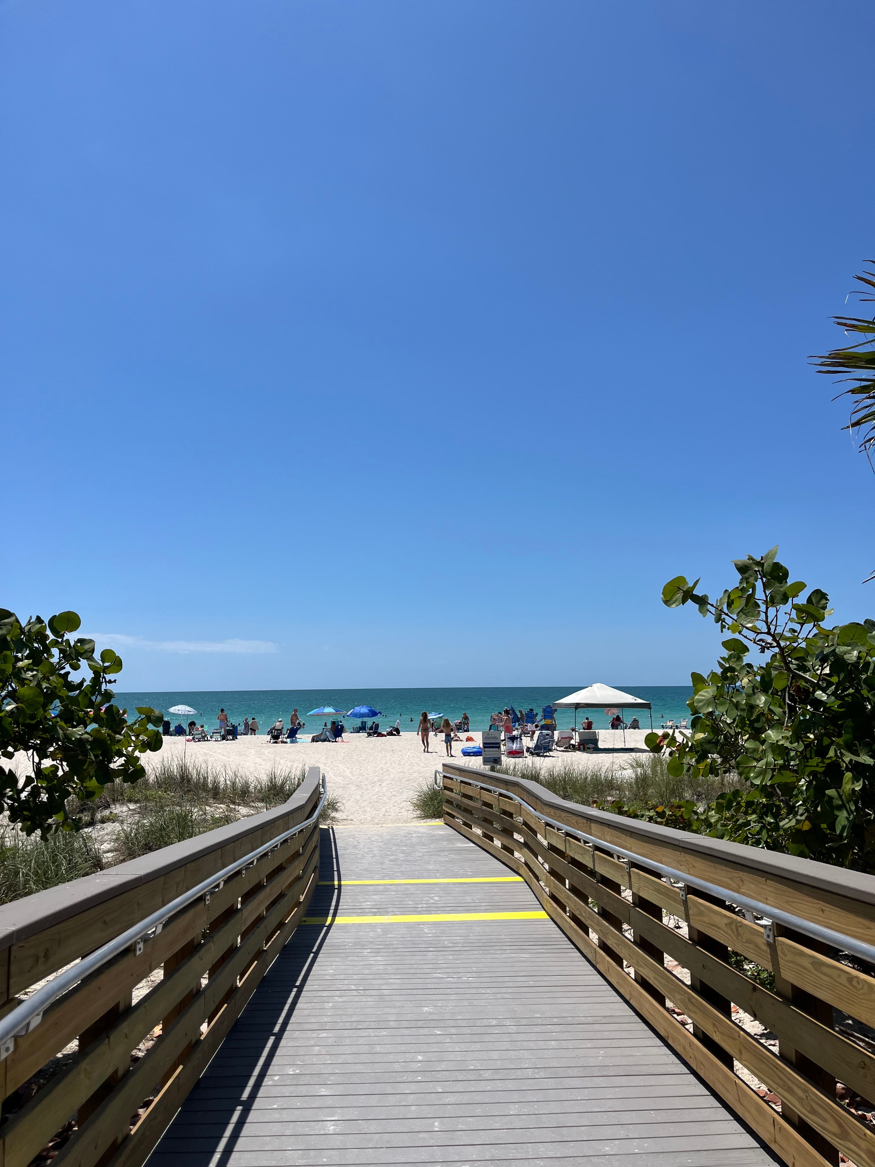 View of an empty wooden dock leading to a clean beach with lounge chairs on a sunny day