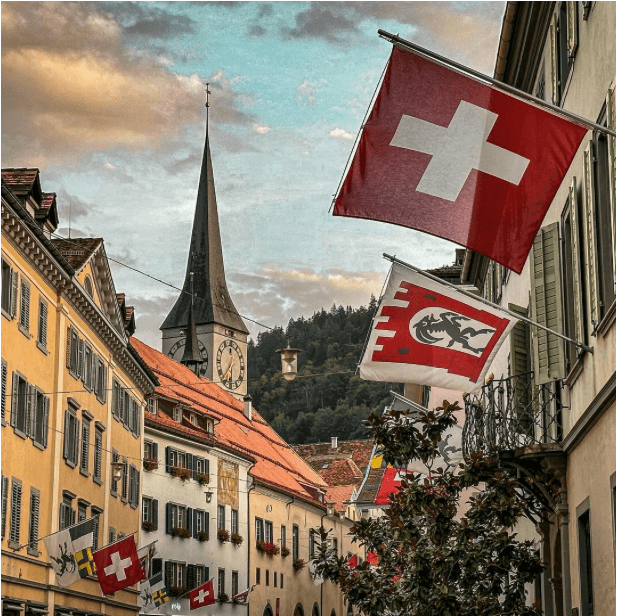A street lined with historical building that have flags in the daytime with a mountain range in the distance.