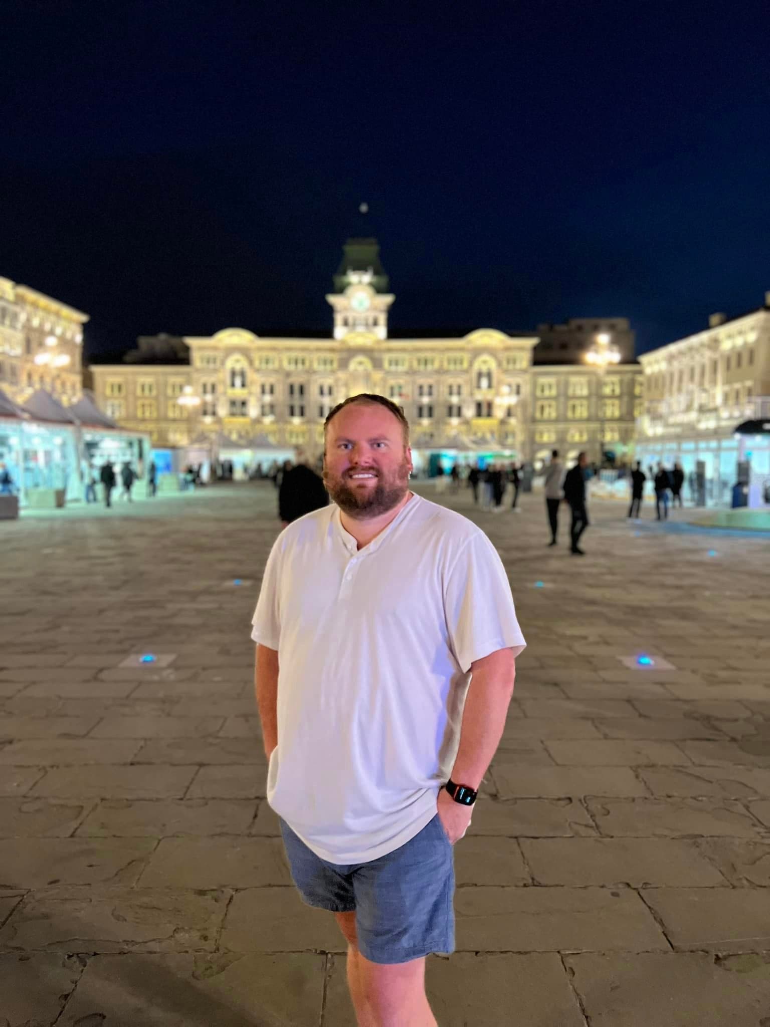 A man standing and smiling in front of a large white historical building at night.