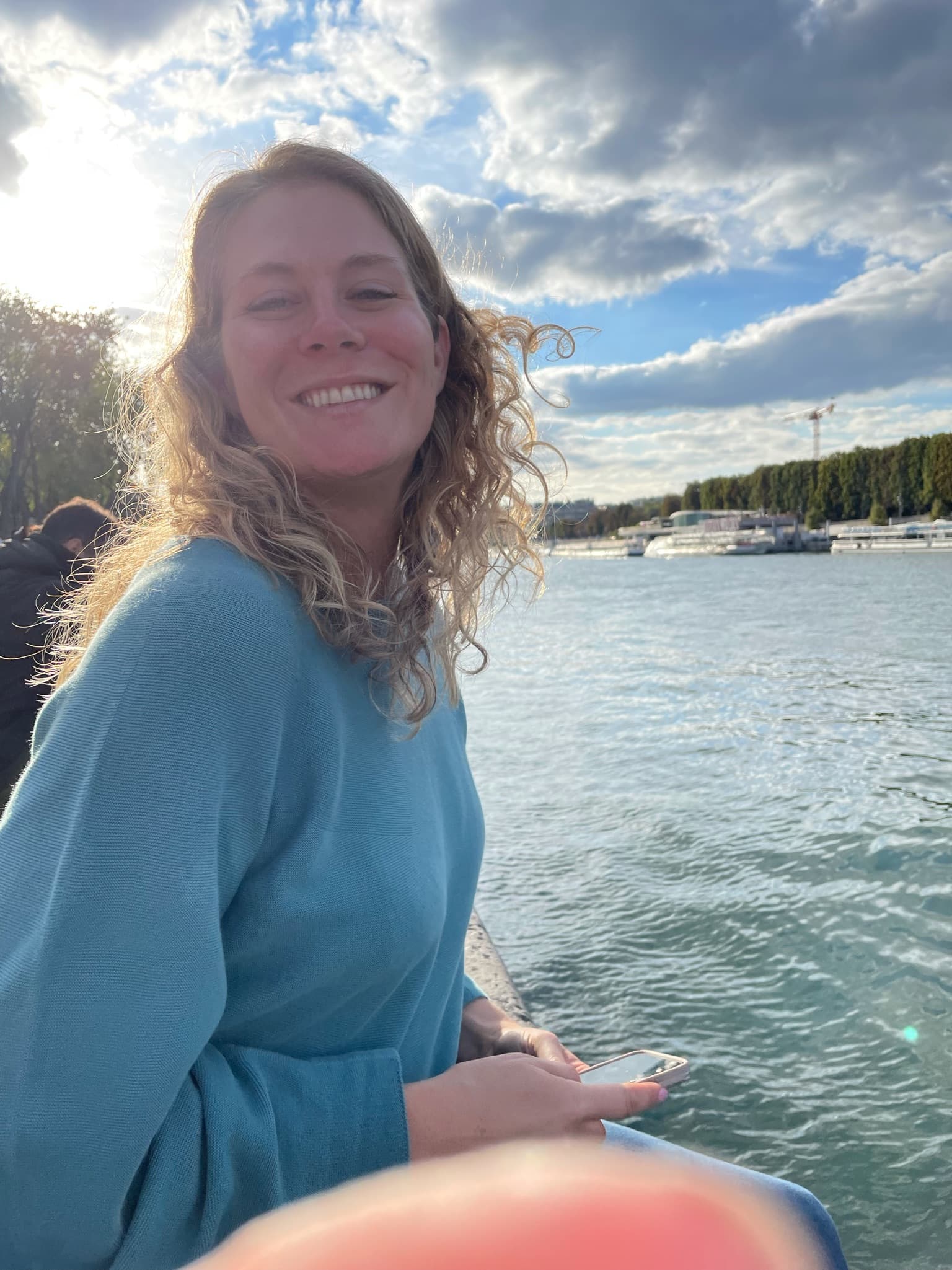 Advisor posing near the water with a boat in the background on a sunny day with clouds parting above. 