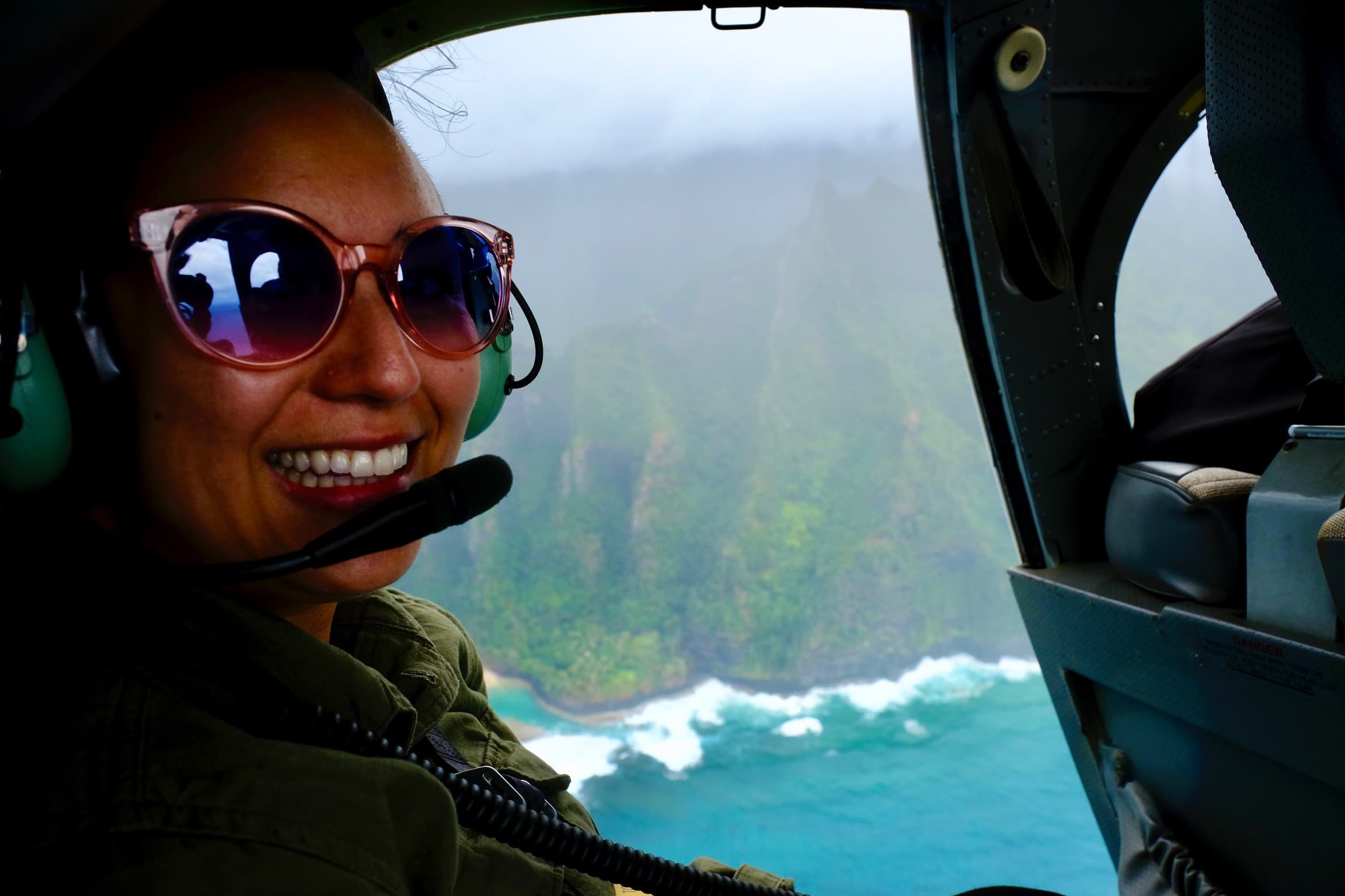 A person smiling with a pilots headset on, flying over a body of water.  Waves crashing on the beach and forest in the distance.