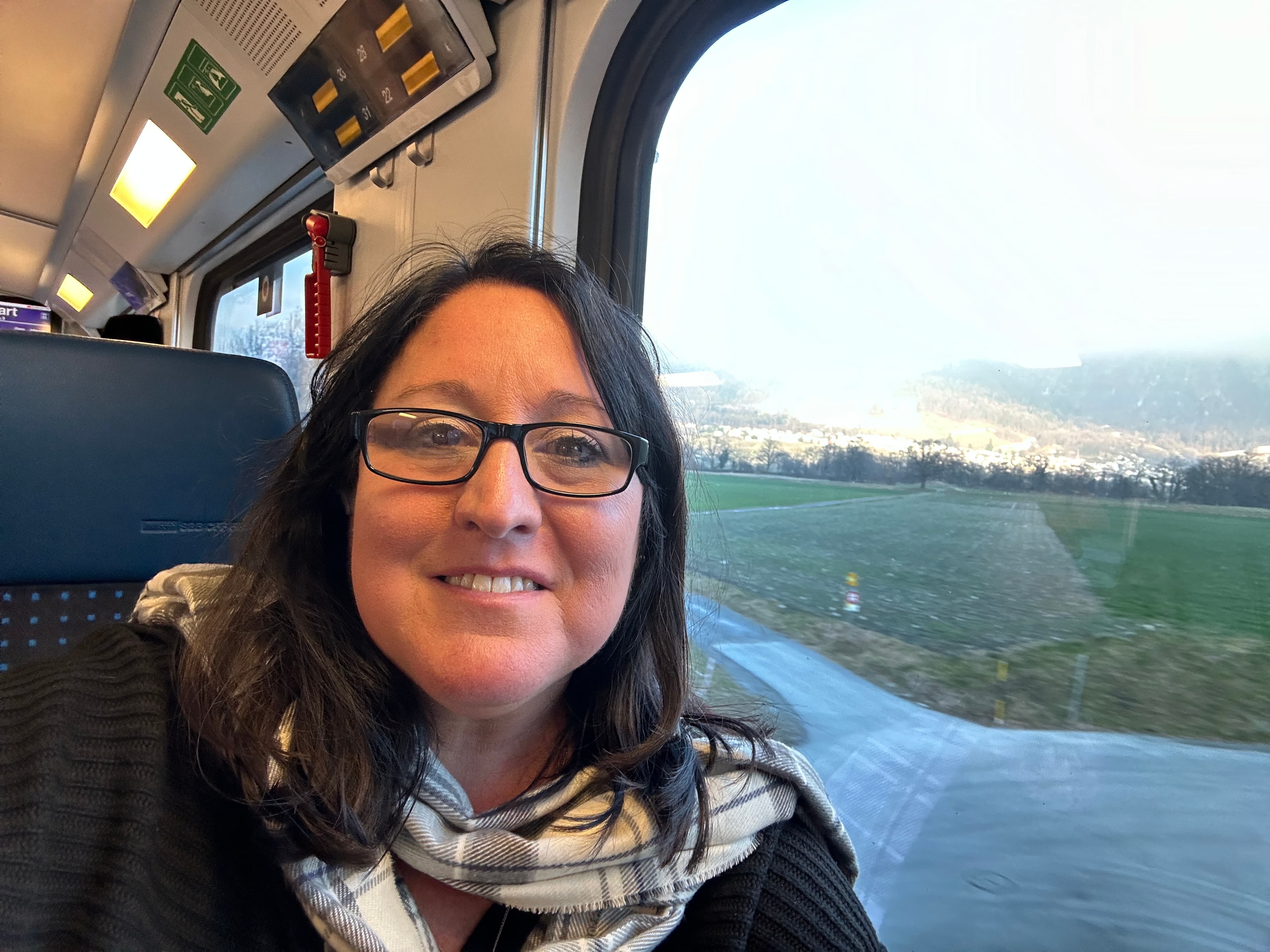 A person posing for a selfie inside of a train with a view of the countryside outside of the window. 