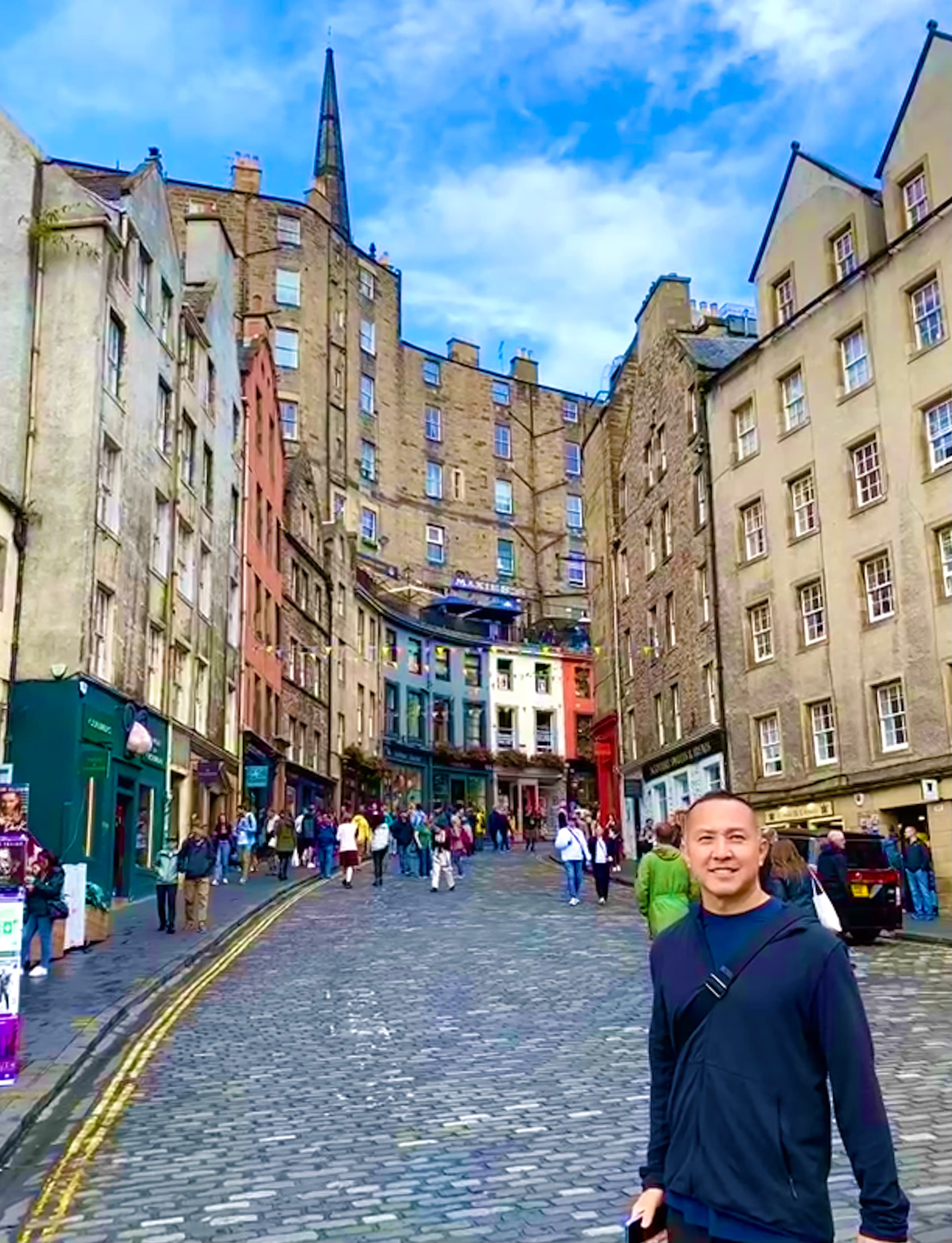 Scott in a black shirt posing on an old stone street lined with pretty buildings and some pedestrians on a sunny day
