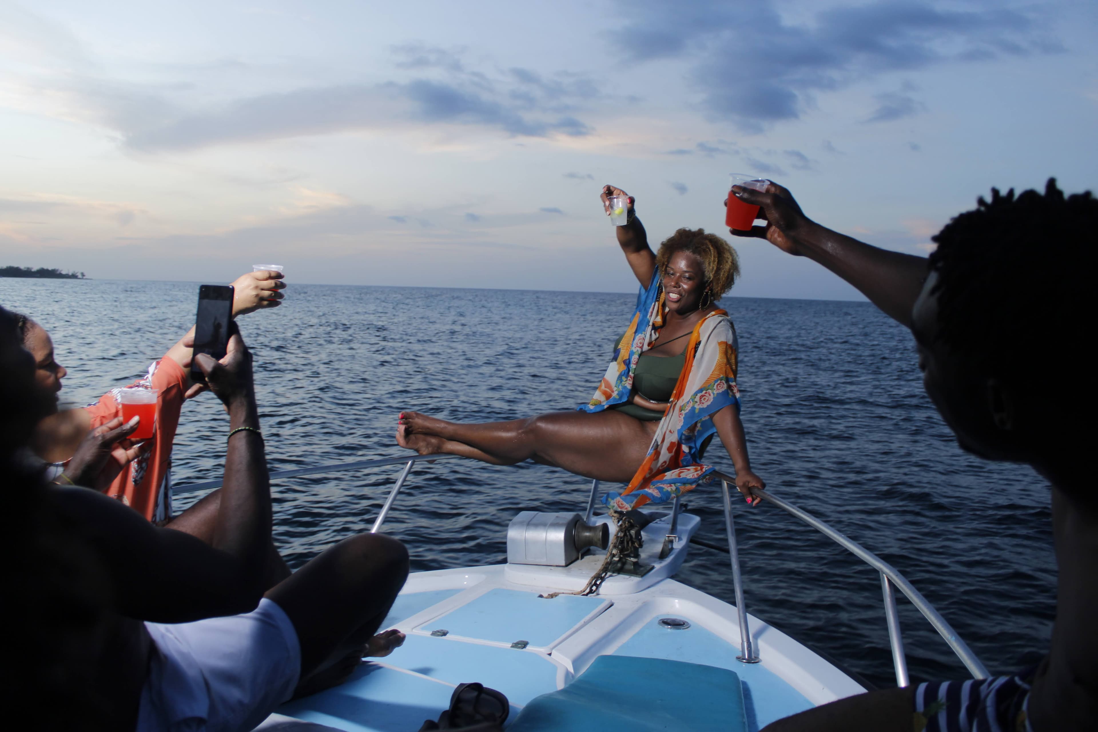 A woman sitting on the bow of a boat with her glass raised in a toast with friends surrounding her at sunset.