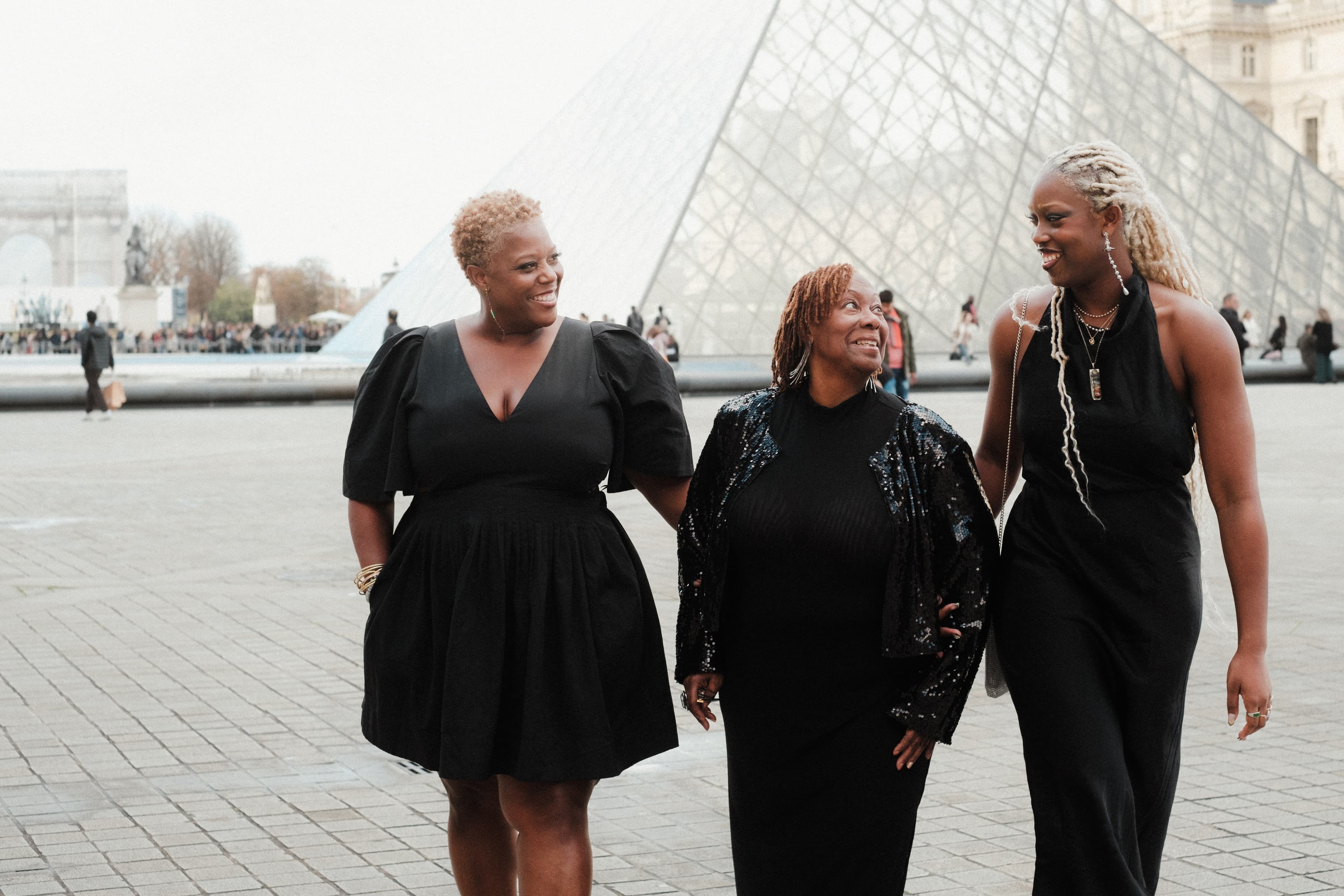 Three women walking together in front of a large pyramid-shaped glass sculpture during the day.