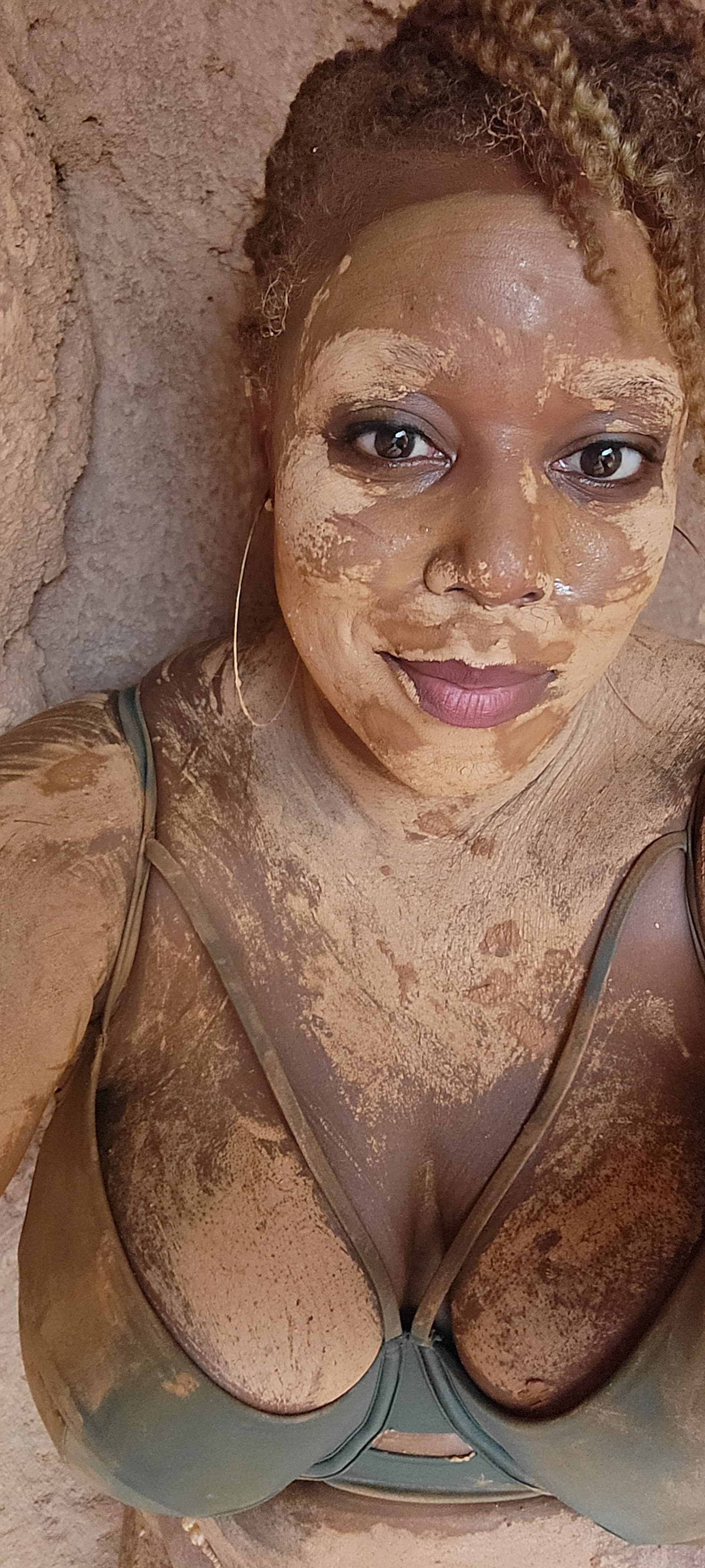 A woman covered in a mud mask relaxing against a stone wall.