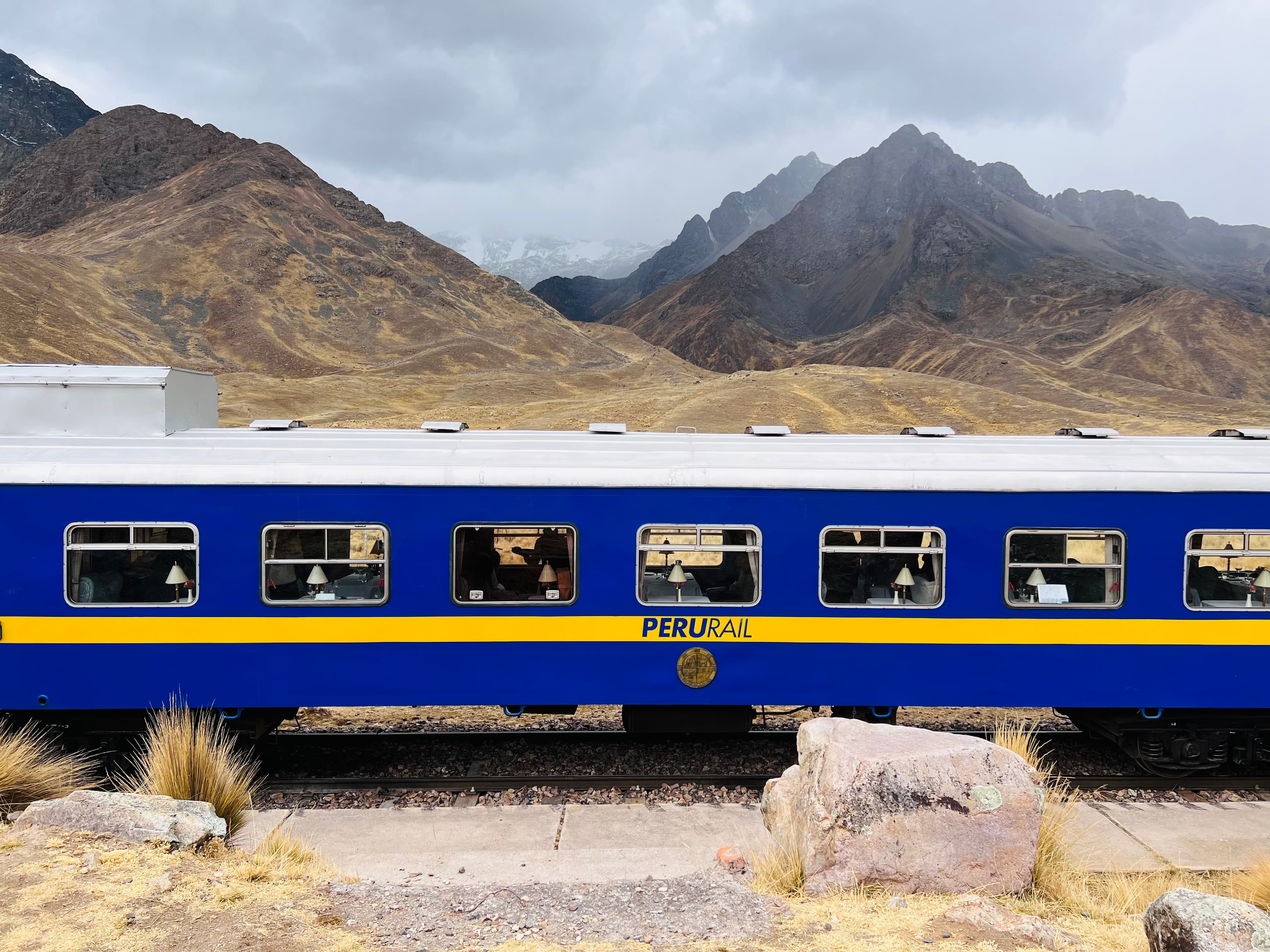 Photo of a blue train with a yellow stripe overlooking mountains in the background.