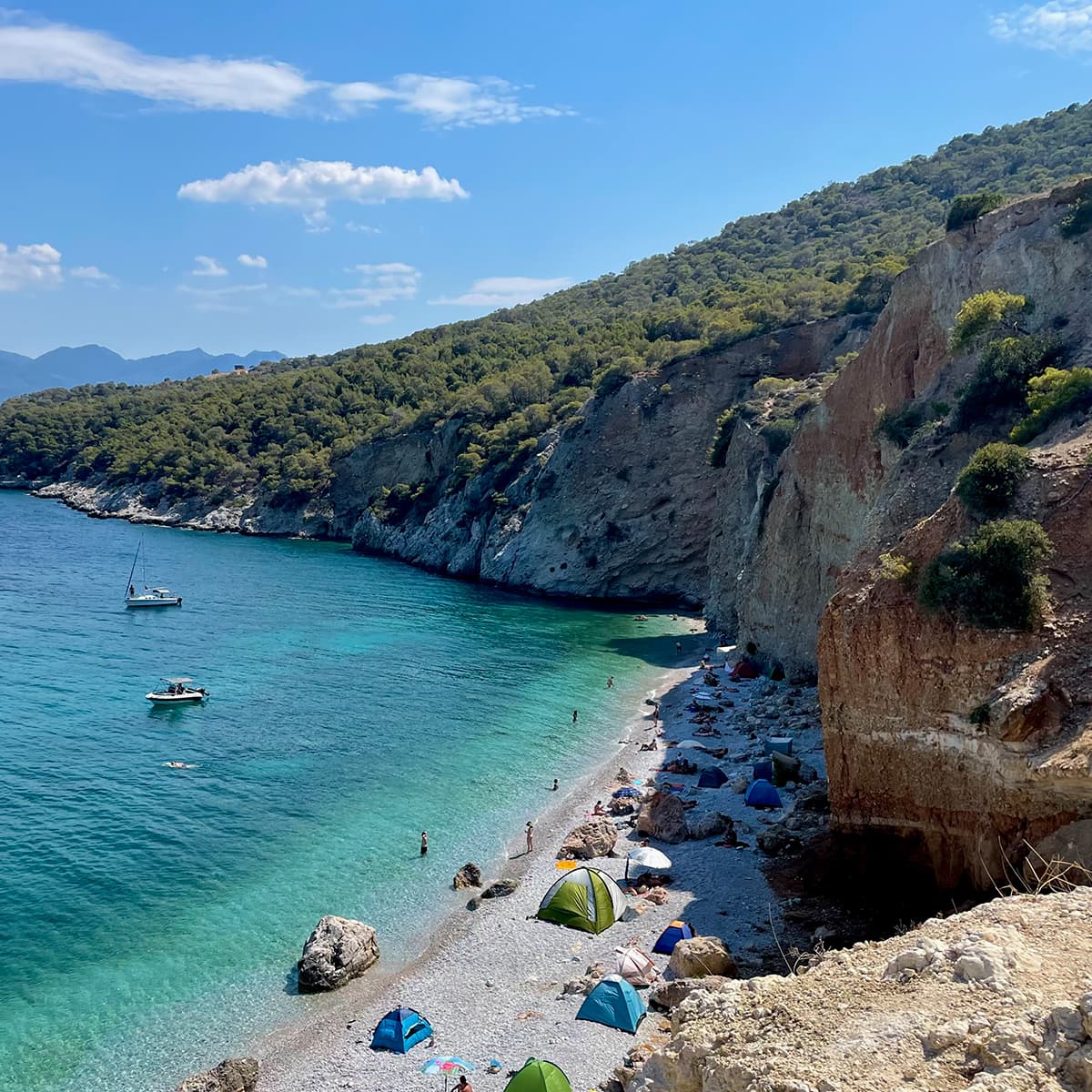 Beautiful aerial view of a pebble beach with sunbathers and a turquoise ocean next to rocky cliffs on a sunny day