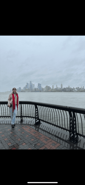 A person standing against a fence overlooking the ocean with a city skyline and fog in the distance.