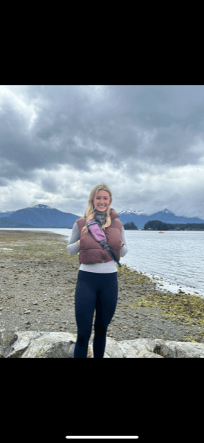 A blonde woman wearing black pants and a mauve colored vest standing on a beach with snowy mountains behind her during the day.