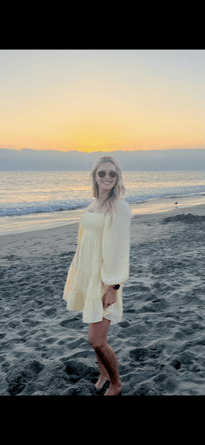 A blonde woman wearing a yellow dress, standing on the beach with the ocean and sunset behind her.