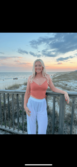 A woman wearing a coral v-neck shirt, standing along a fence overlooking the beach and ocean at sunset.