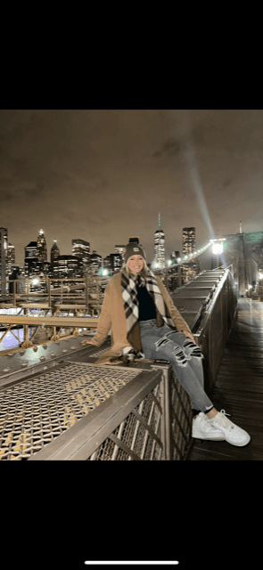 A woman wearing blue jeans, a black beanie, plaid scarf and beige pea coat, sitting on a rooftop with a city skyline behind her at night.