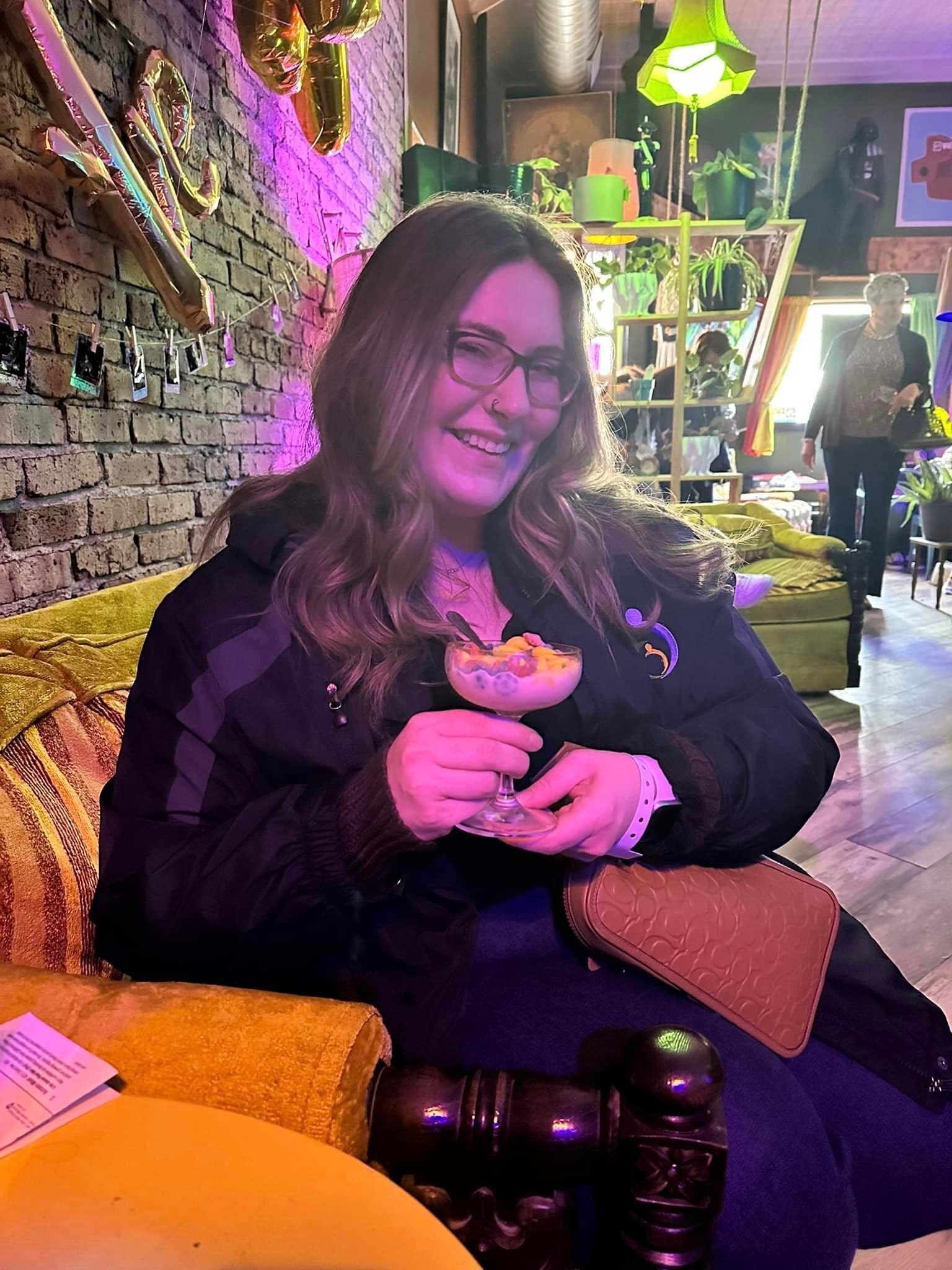 Danica in a black jacket holding a cocktail while sitting on a gold couch indoors against a brick wall