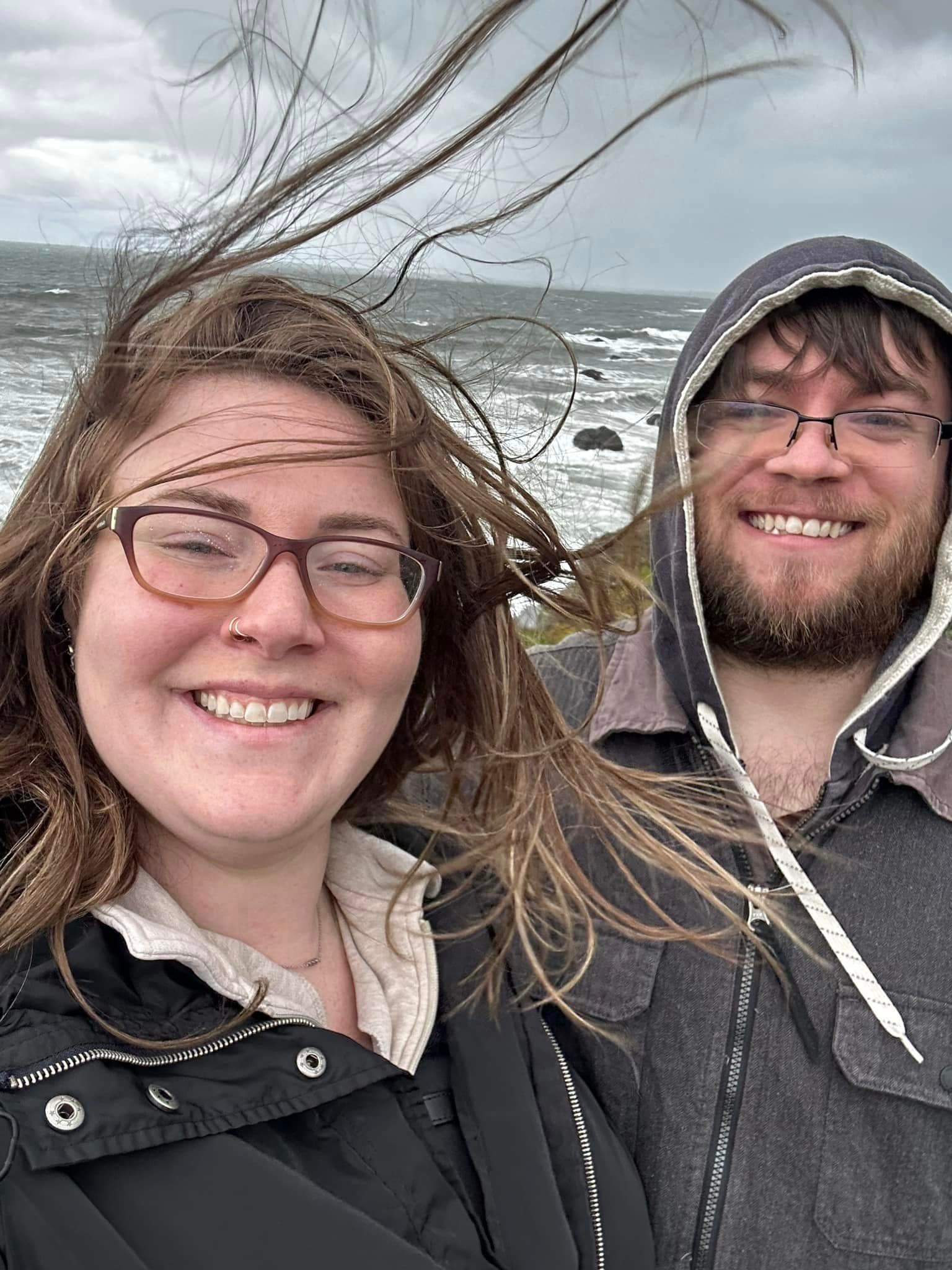 Danica and a man taking a selfie in a windy climate with a grey ocean and cloudy skies behind them