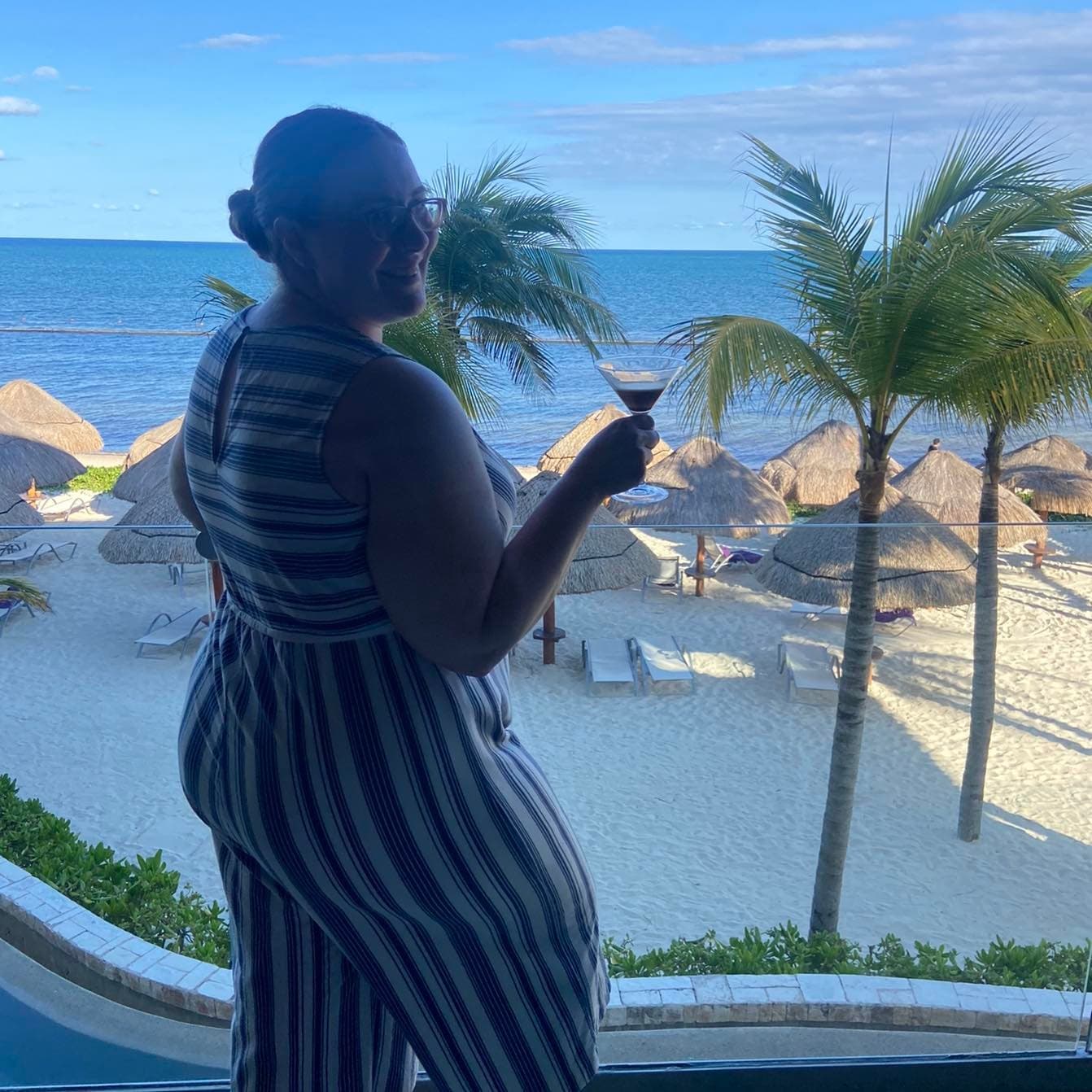 Danica in a striped jumpsuit holding a martini glass overlooking a beautiful beach with lounges and palm trees