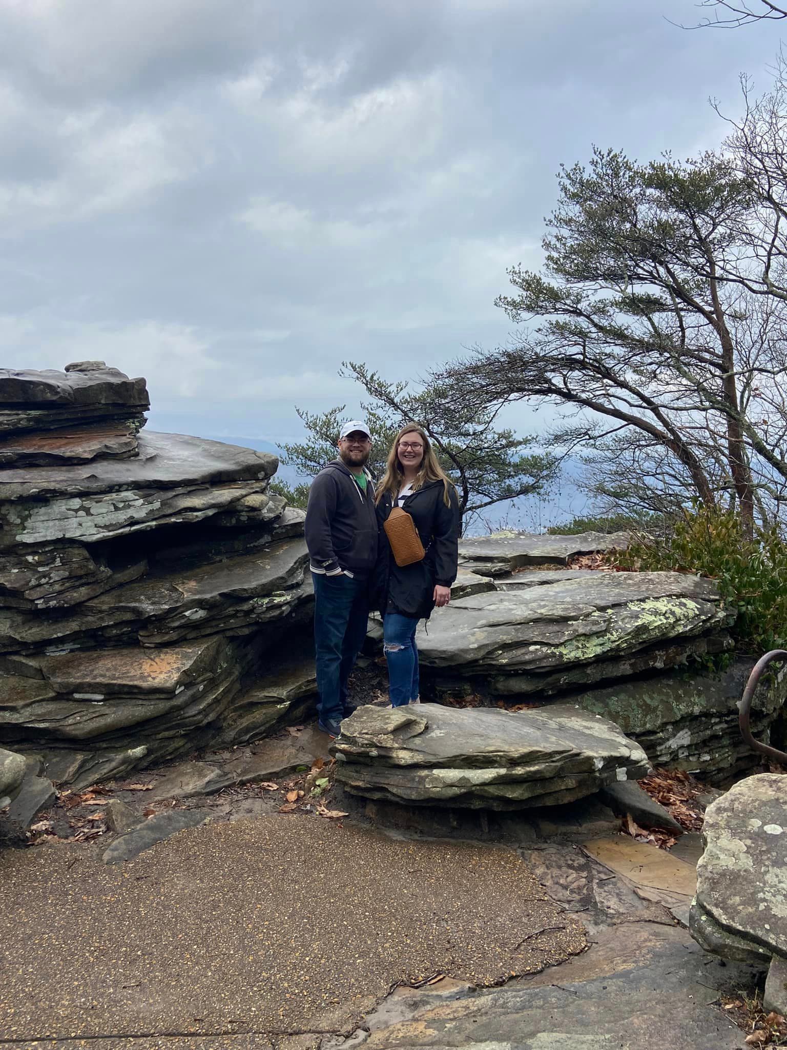 Danica and a man posing side by side on a rock on a cloudy day with the ocean visible behind them