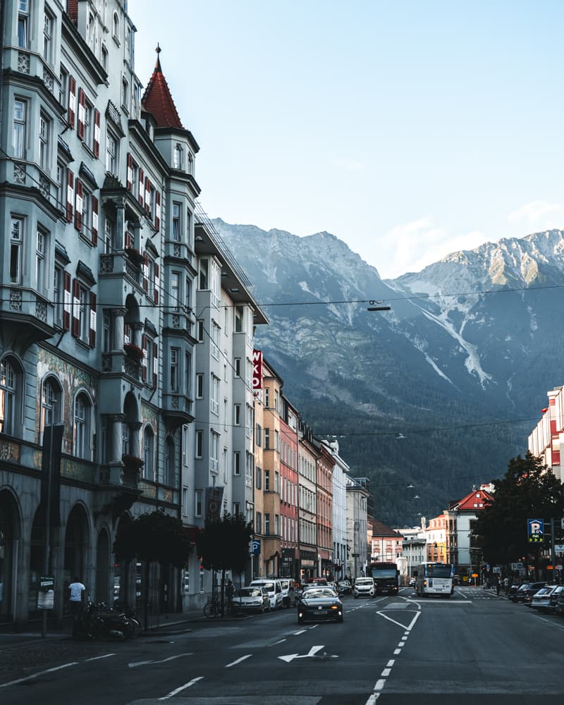 Quaint buildings align under a craggy mountain scene ona clear day dotted with clouds. 