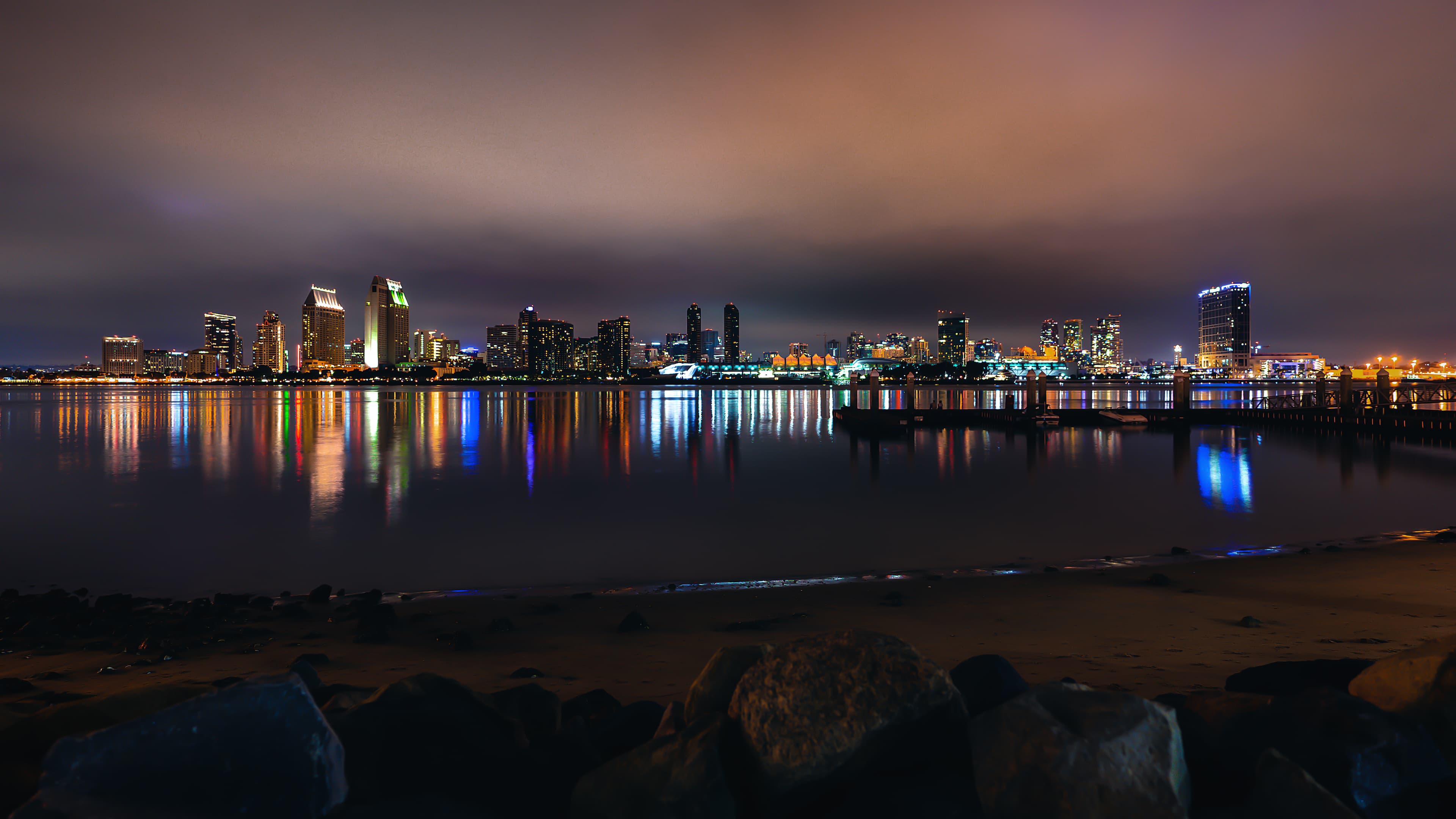 A glowing, neon cityscape lights up the evening sky along a large body of water. 