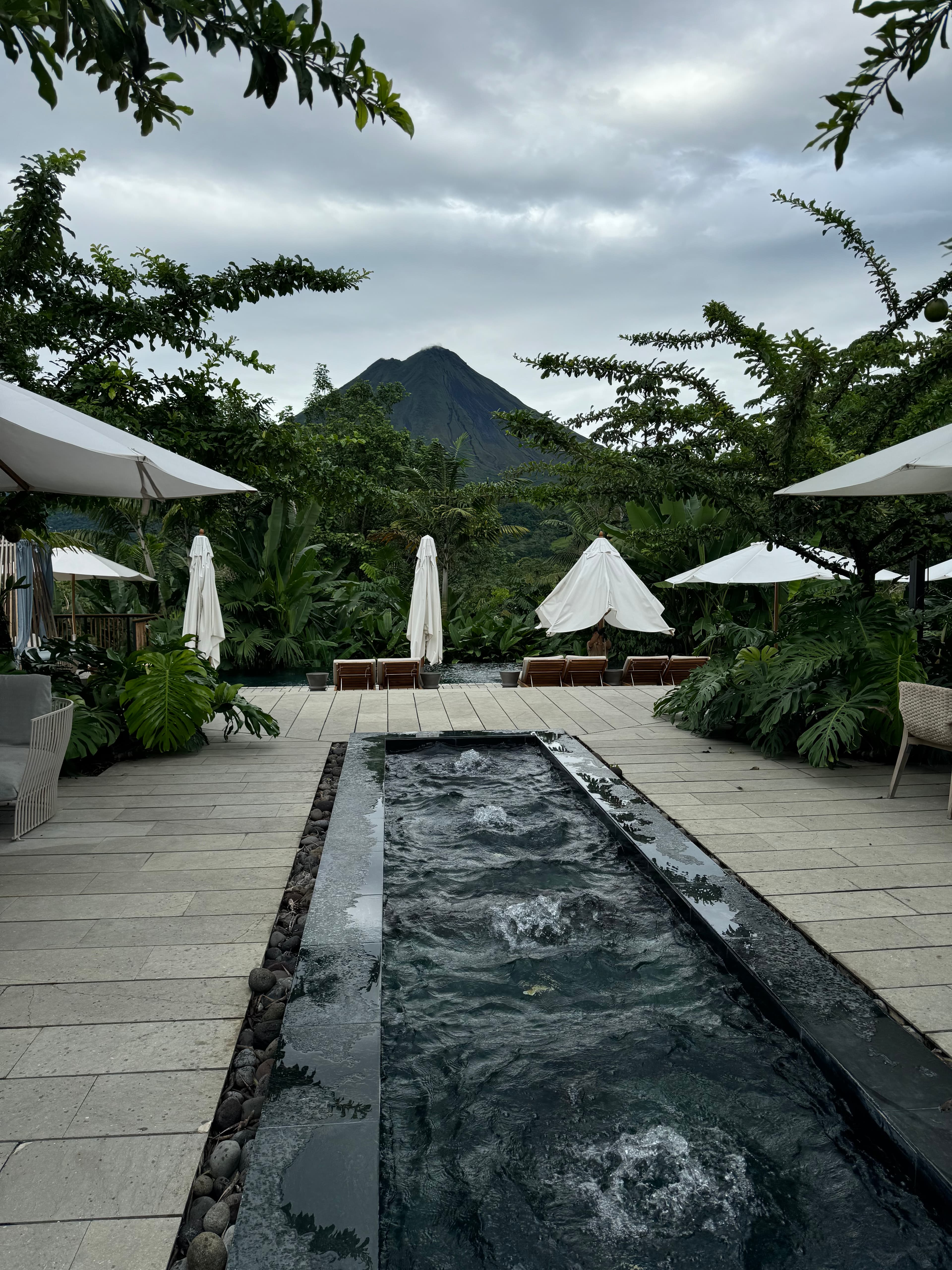 Pretty view of a rectangular fountain surrounded by white umbrellas and green plants with lounge chairs behind it on a cloudy day
