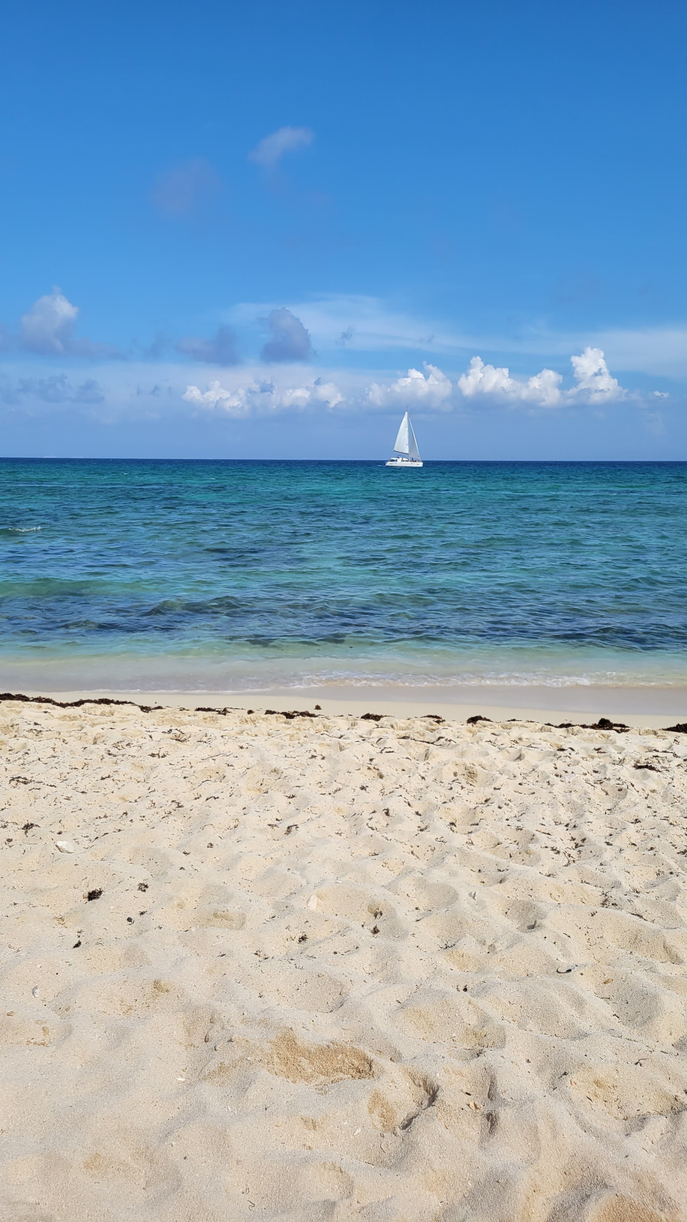 View of an empty white sand beach and the sea during the day with a single sailboat seen on the horizon