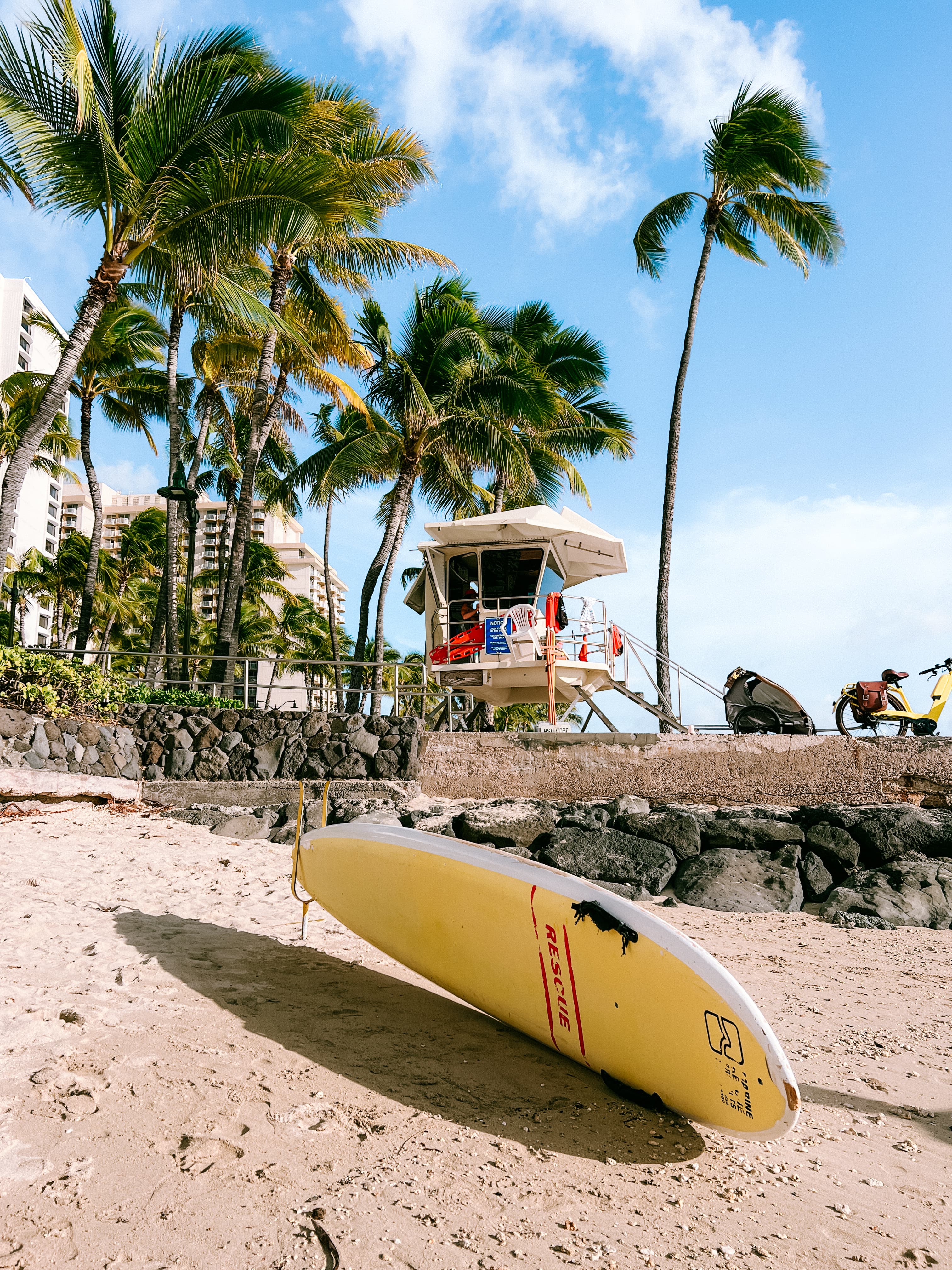 View of a surfboard on a beach with a lifeguard tower and palm trees behind it on a sunny day
