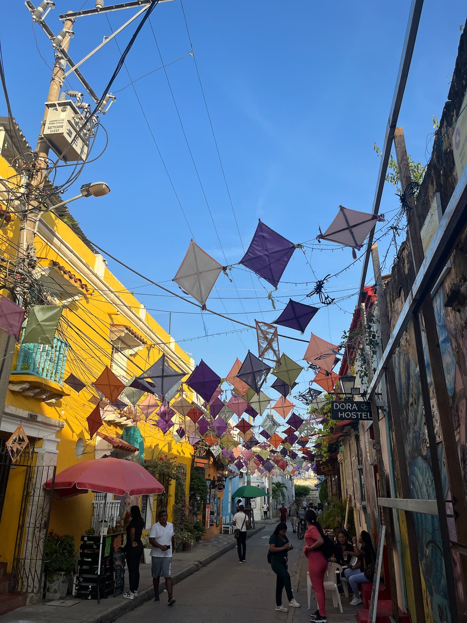 Colorful street scene with kites hanging overhead, vibrant buildings, and people walking along the street.
