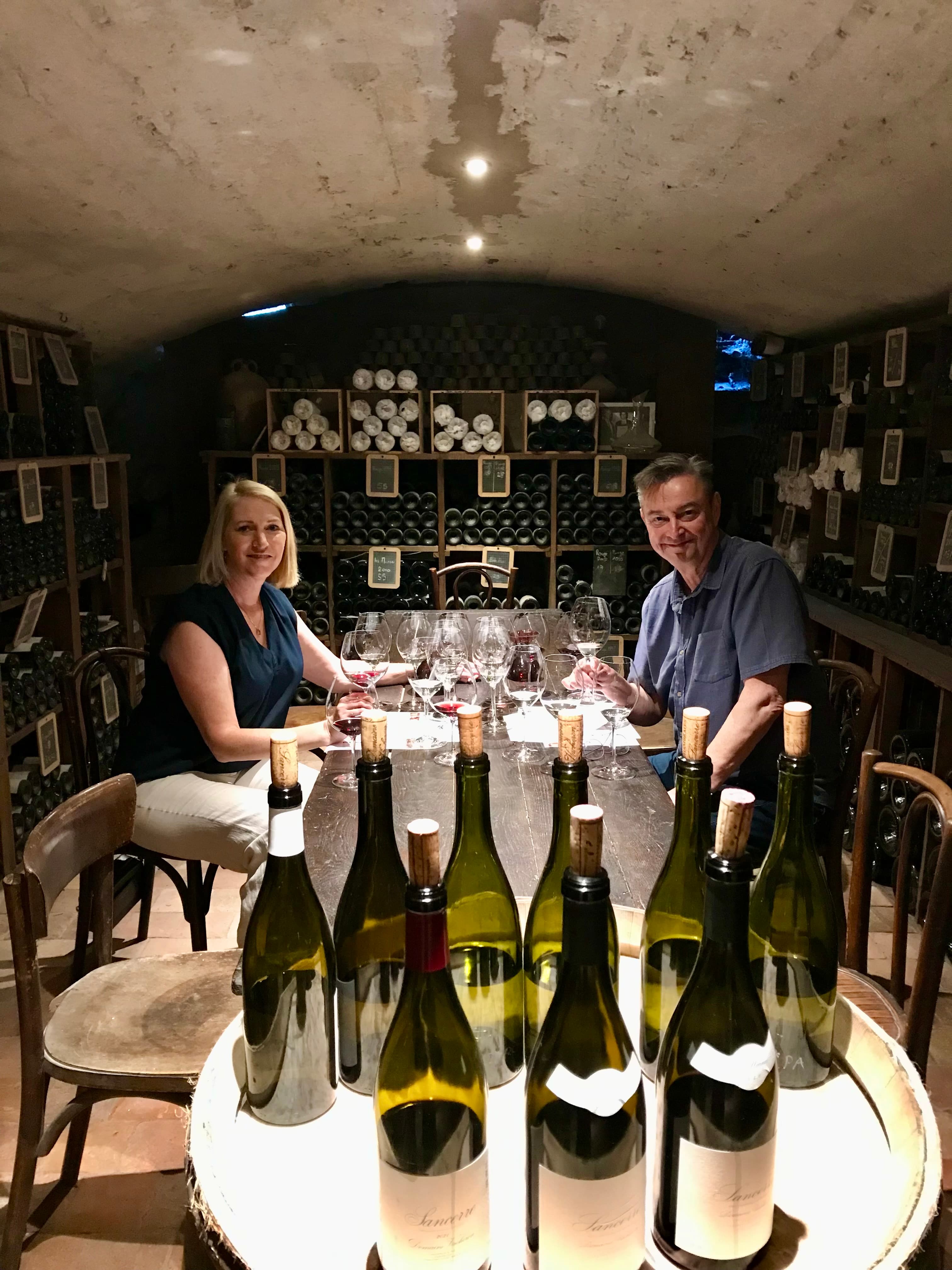 Leslie and her husband seated at a table in a wine cellar with many bottles of wine around them
