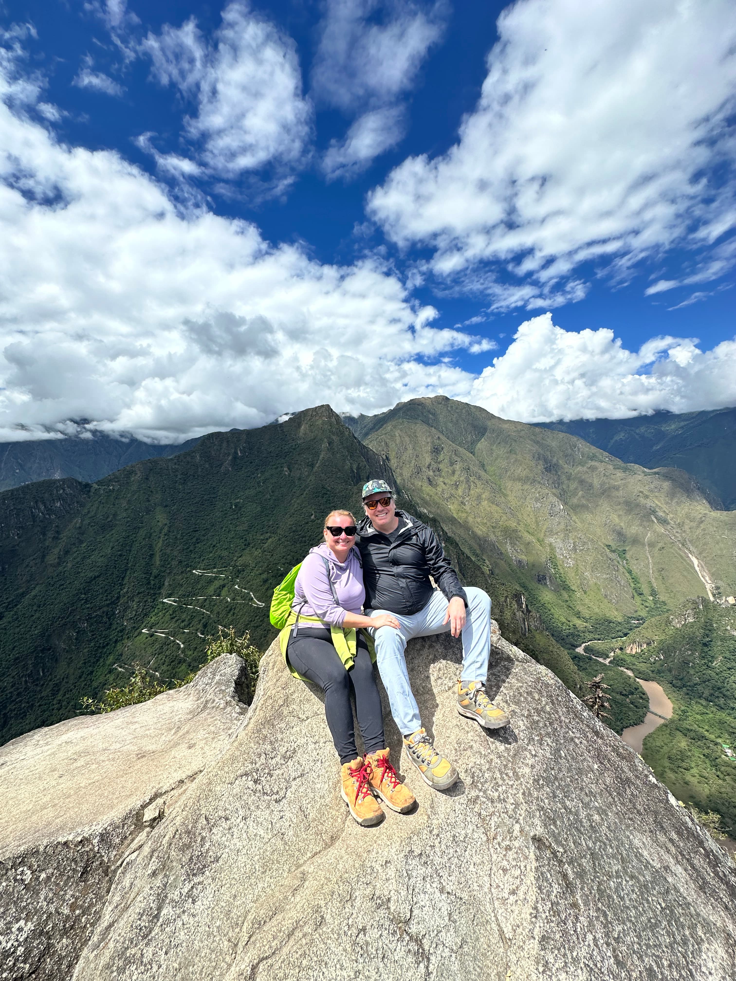 A couple posing for an image at the top on a hiking range on a sunny day. 