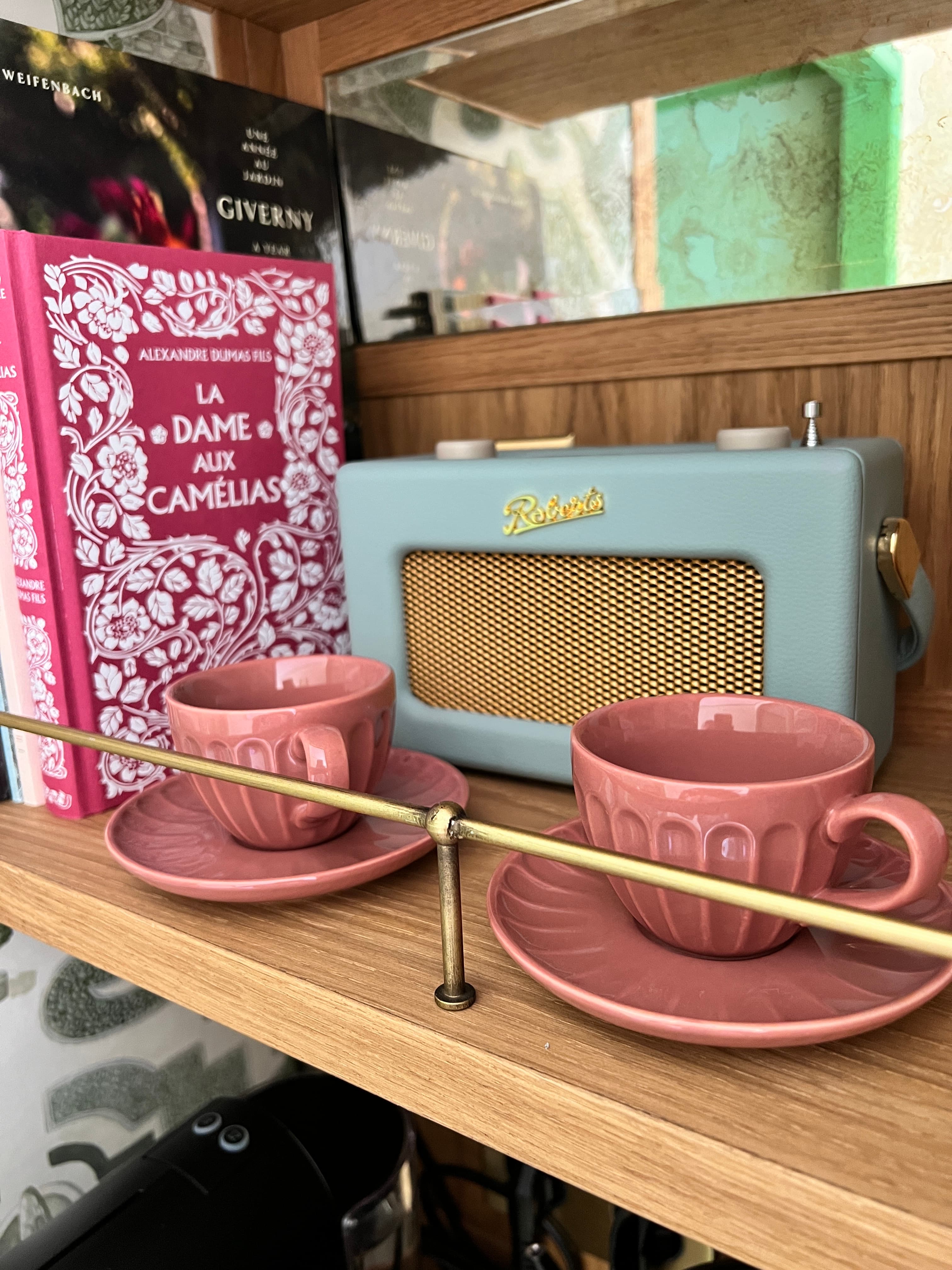 View of two pink teacups and matching saucers on a wooden shelf next to a blue speaker
