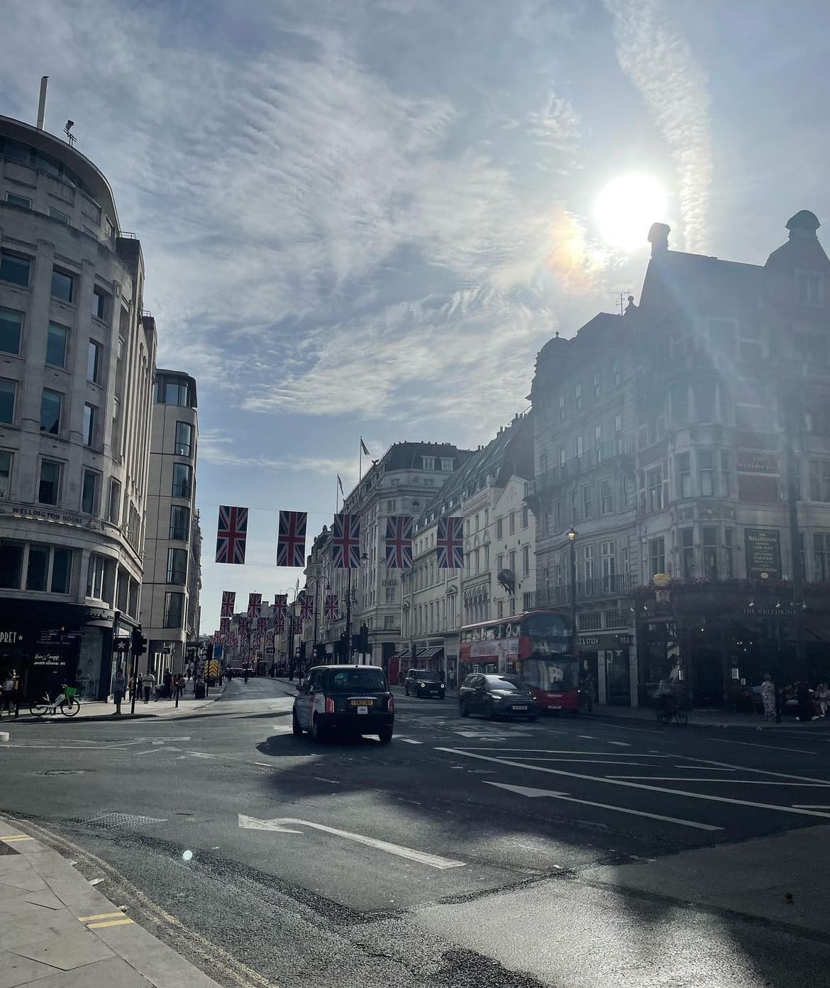 Sun light shines down on a busy street corner lined with tall, historic buildings under a cloudy sky. 