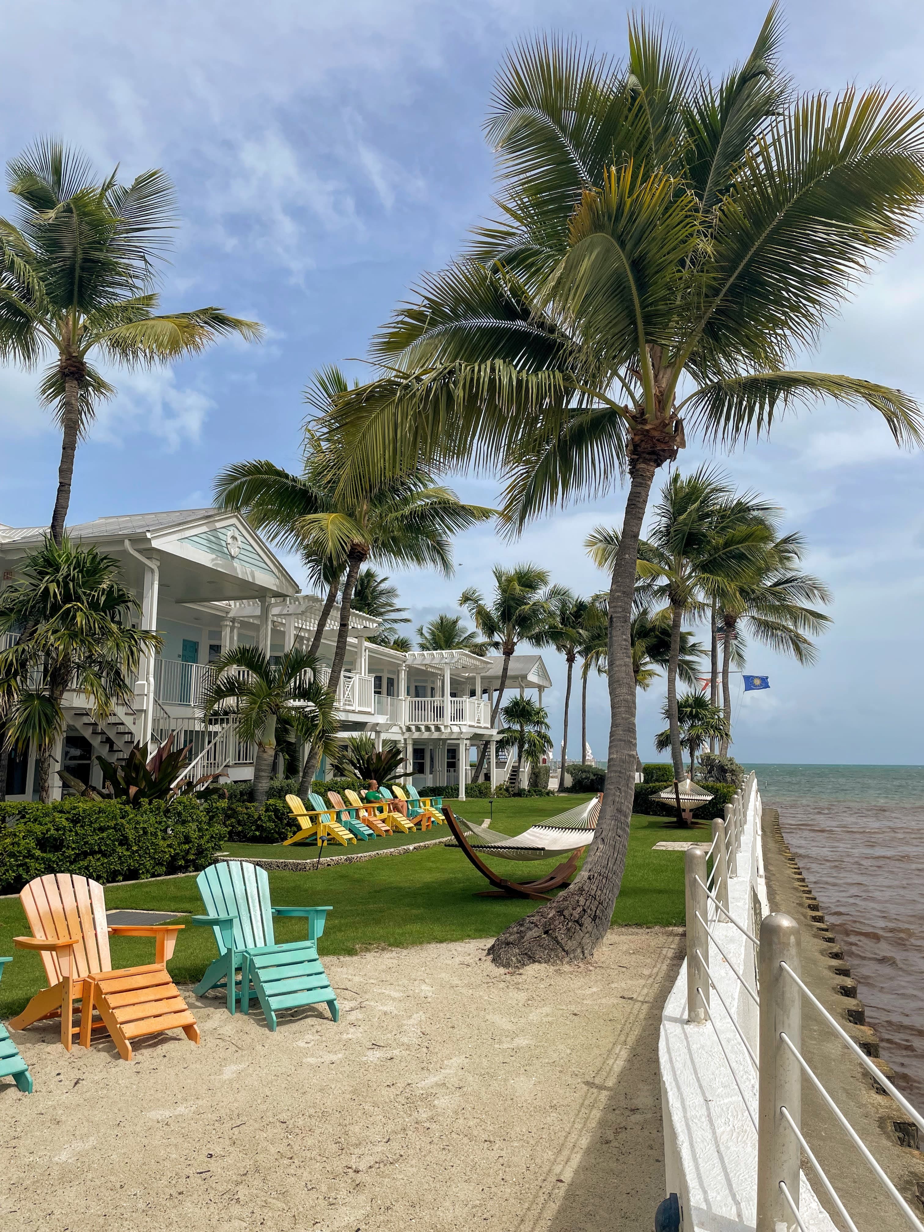 View of two colorful Adirondack chairs on a deck with palm trees and hammocks overlooking the sea