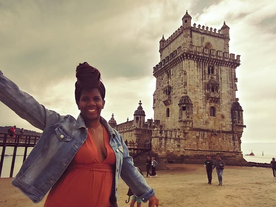 A woman poses in front of a tower, wearing an orange shirt and jeans.