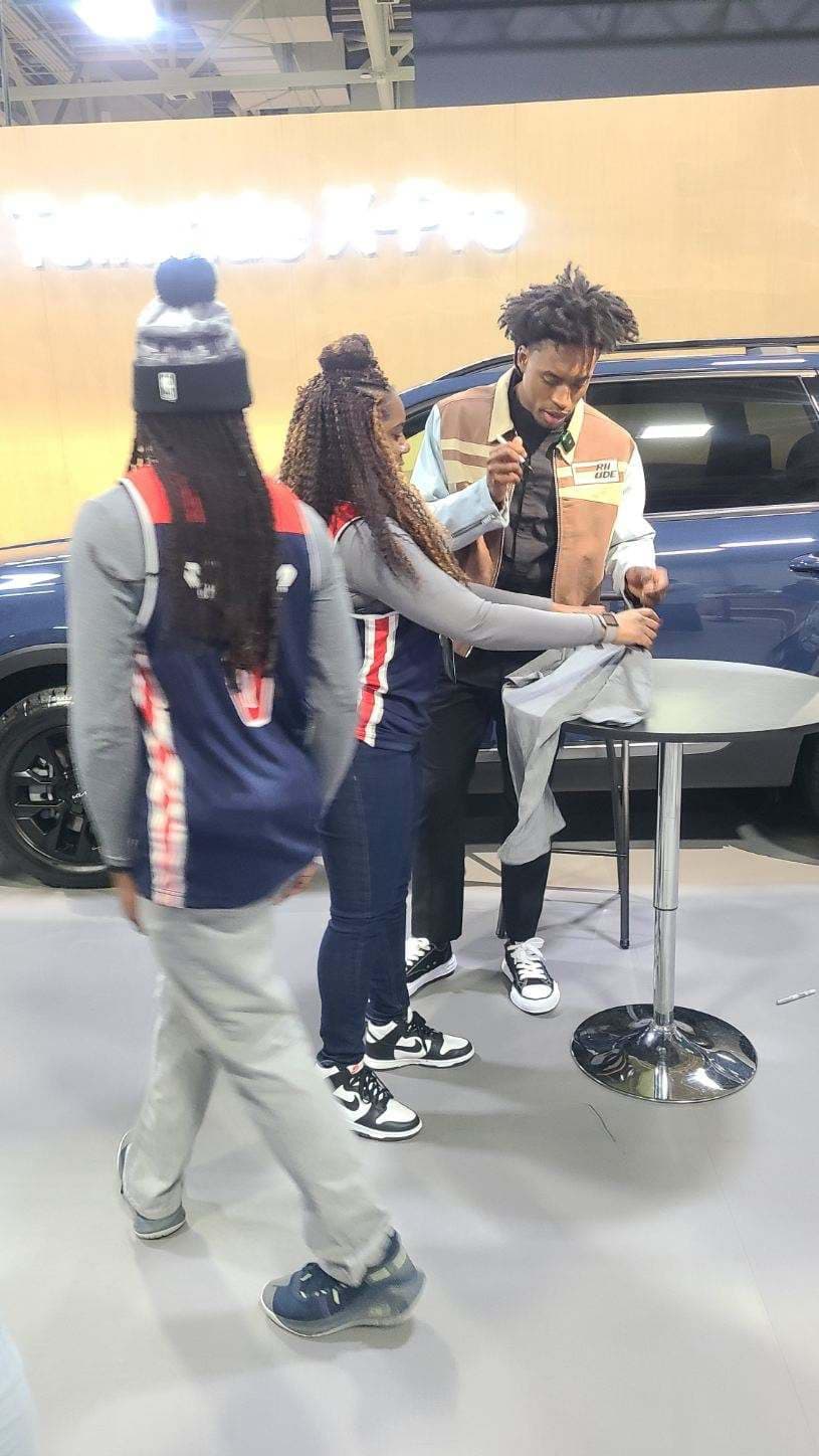Three people at a table, with one signing an autograph. Casual attire, including a jacket and sneakers.